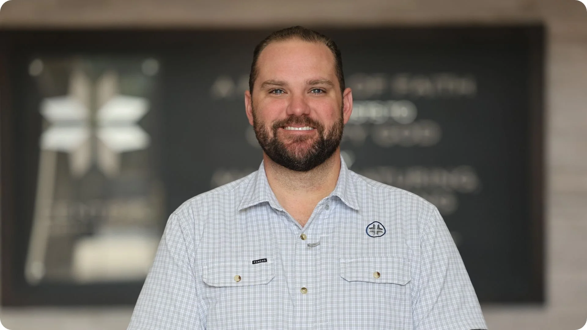 A smiling man with a beard and short hair wearing a blue and white checkered shirt, standing in front of a dark background with a blurred logo or text.