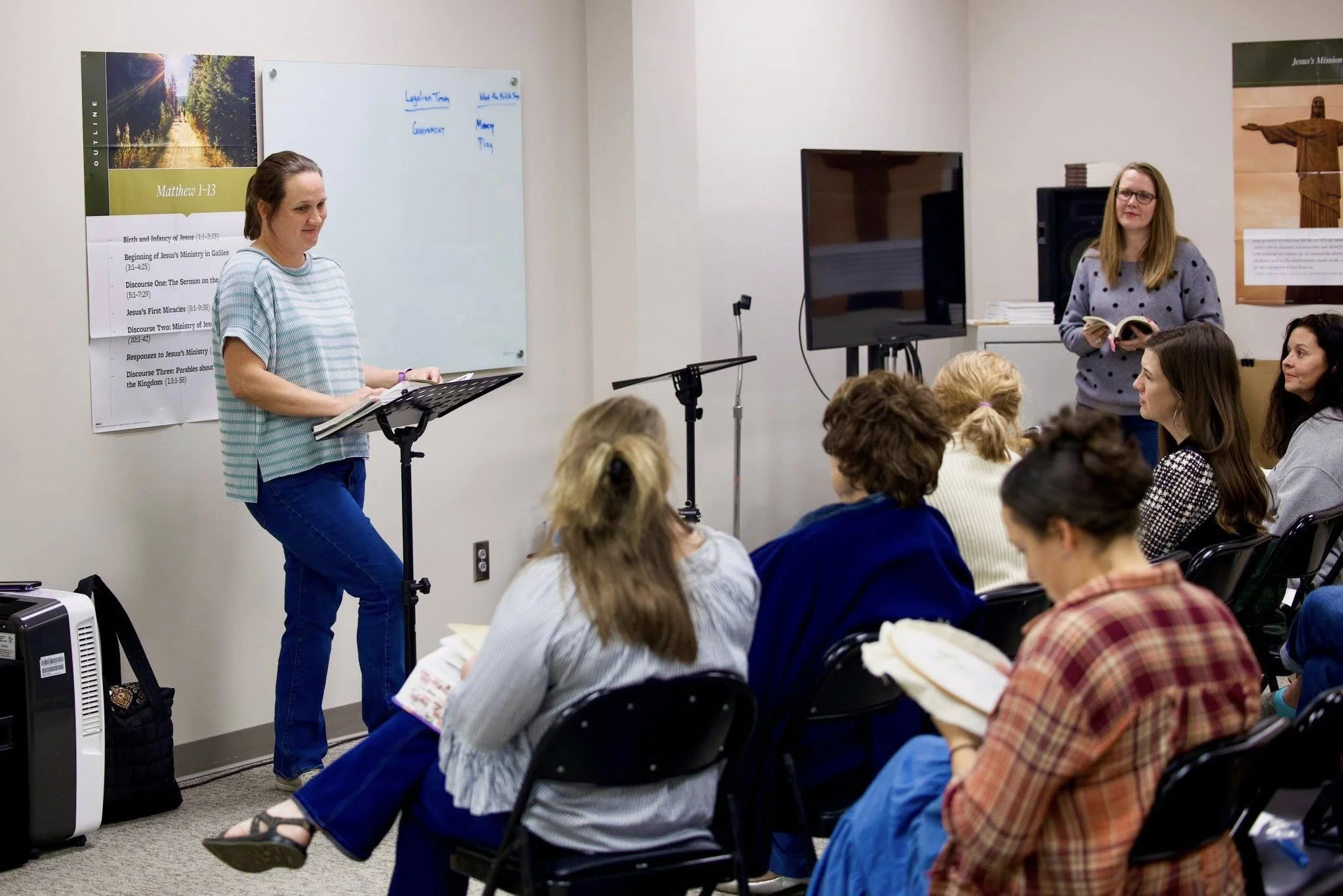 A woman stands at a music stand leading a church discussion or lecture with several people sitting in chairs listening. There is a whiteboard on the wall with notes and a poster about Matthew 1-13 in the background.
