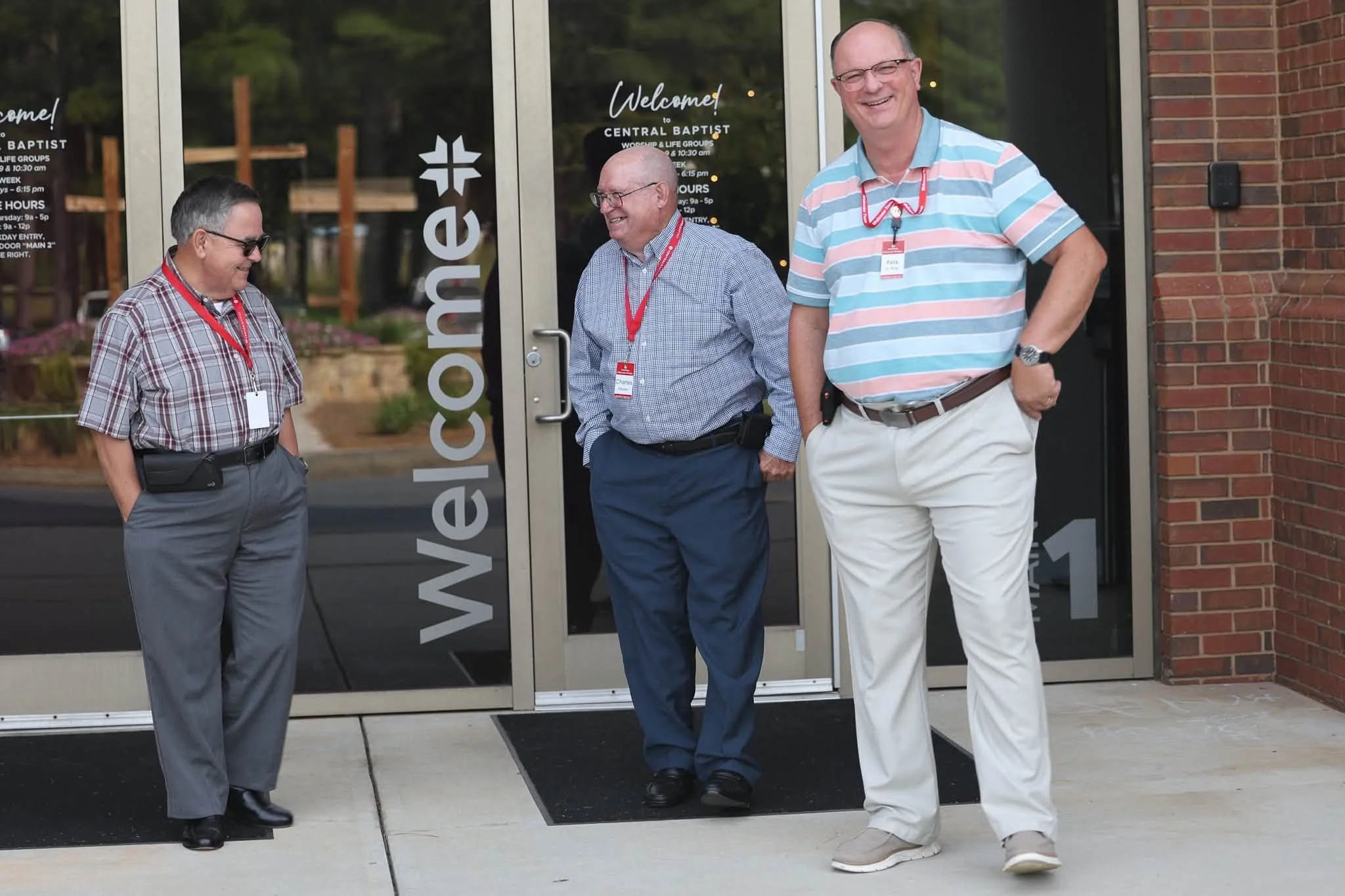 Three men standing outside the entrance of a church, smiling and talking. Two are wearing glasses, and all are dressed in casual business attire with name tags, near glass doors with the church's name and hours listed.