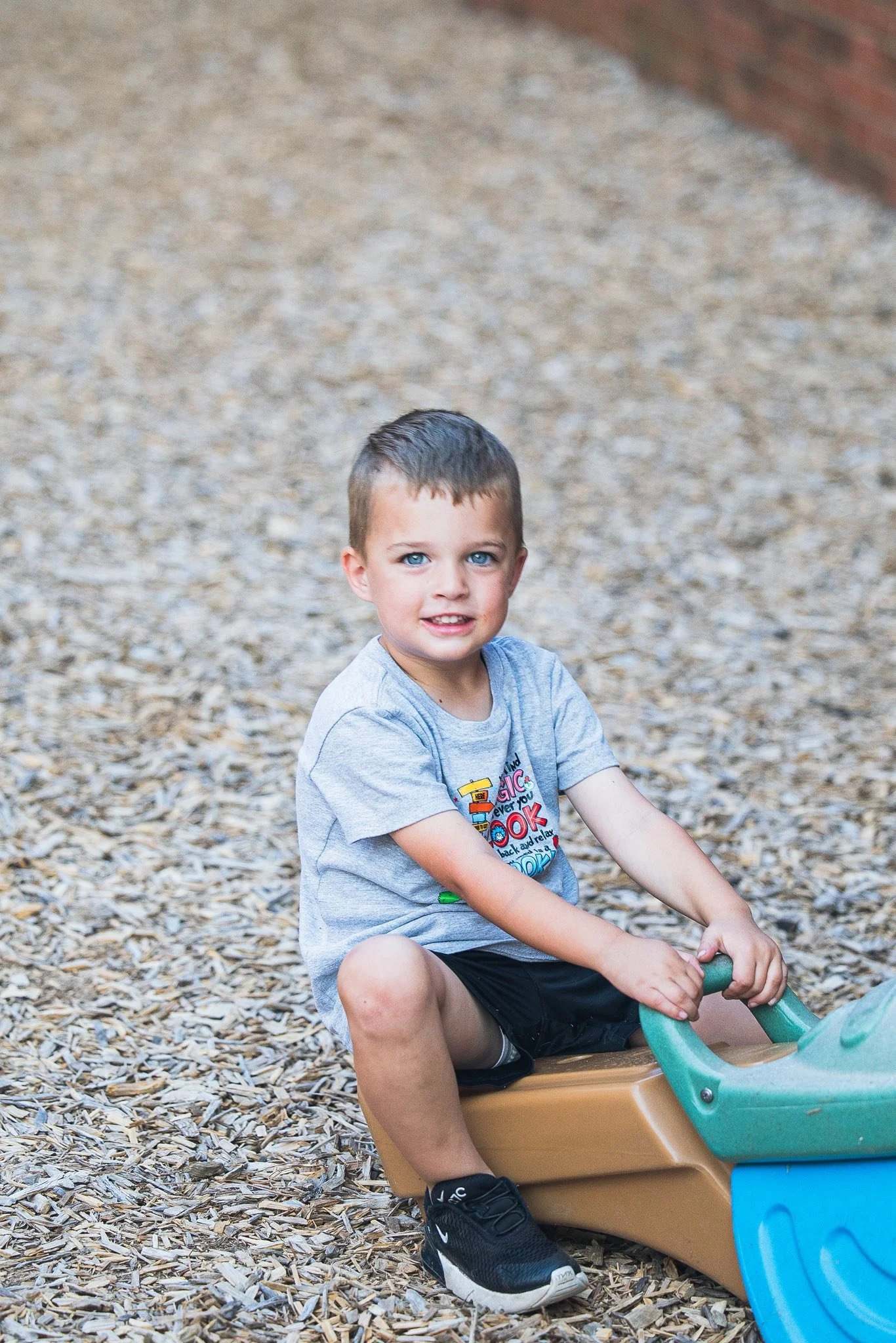 A young boy with blue eyes and short brown hair sitting on a playground slide, holding the slide's handle, surrounded by wood chips, with a brick wall in the background.