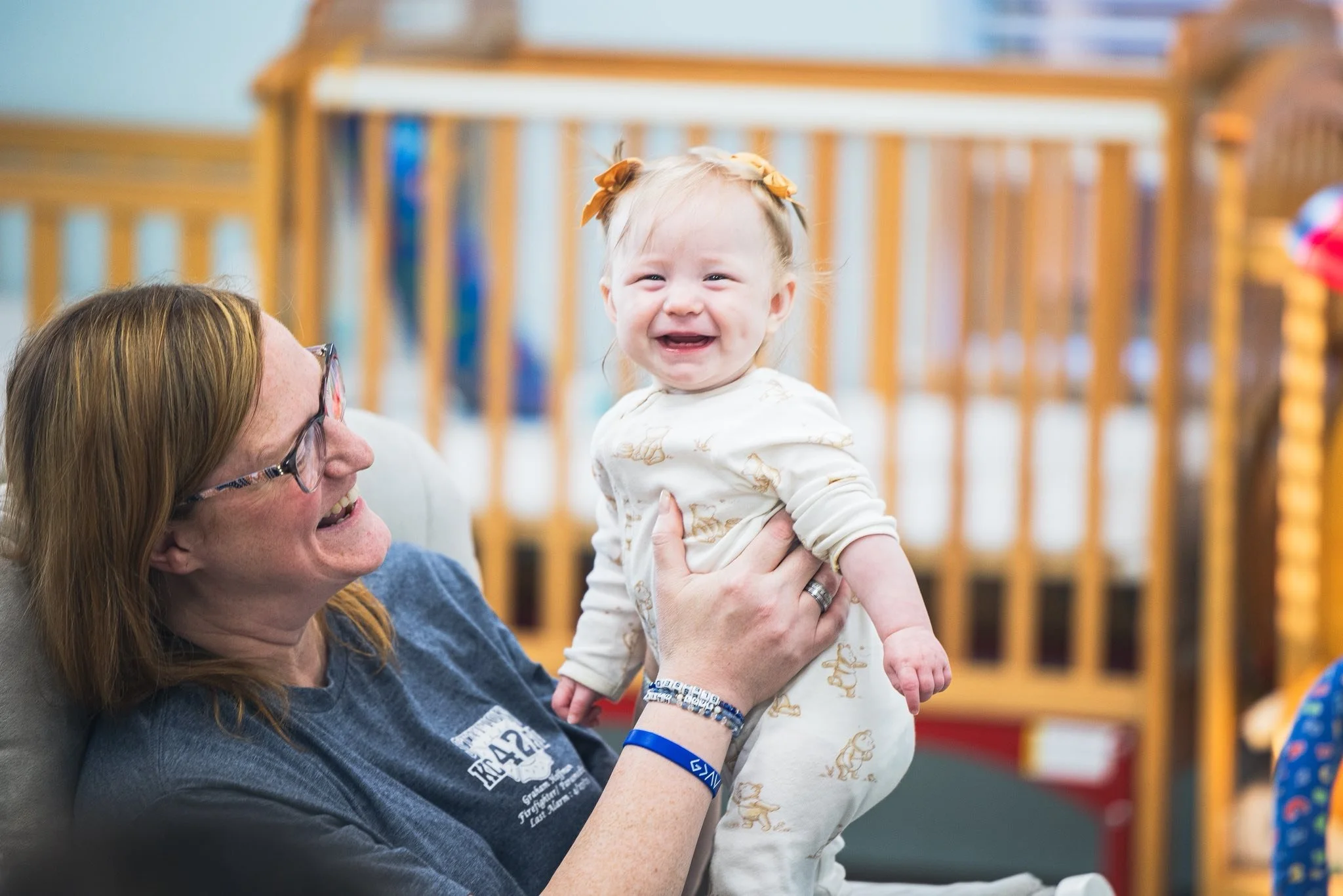 An older woman with red hair and glasses holding a laughing toddler with blonde hair and ribbons, in an indoor setting with wooden railings in the background.