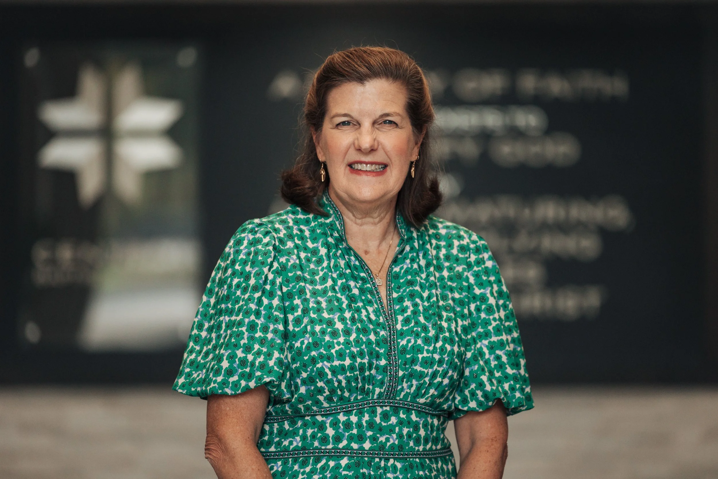 A smiling middle-aged woman with shoulder-length brown hair standing indoors in front of a blurred background.