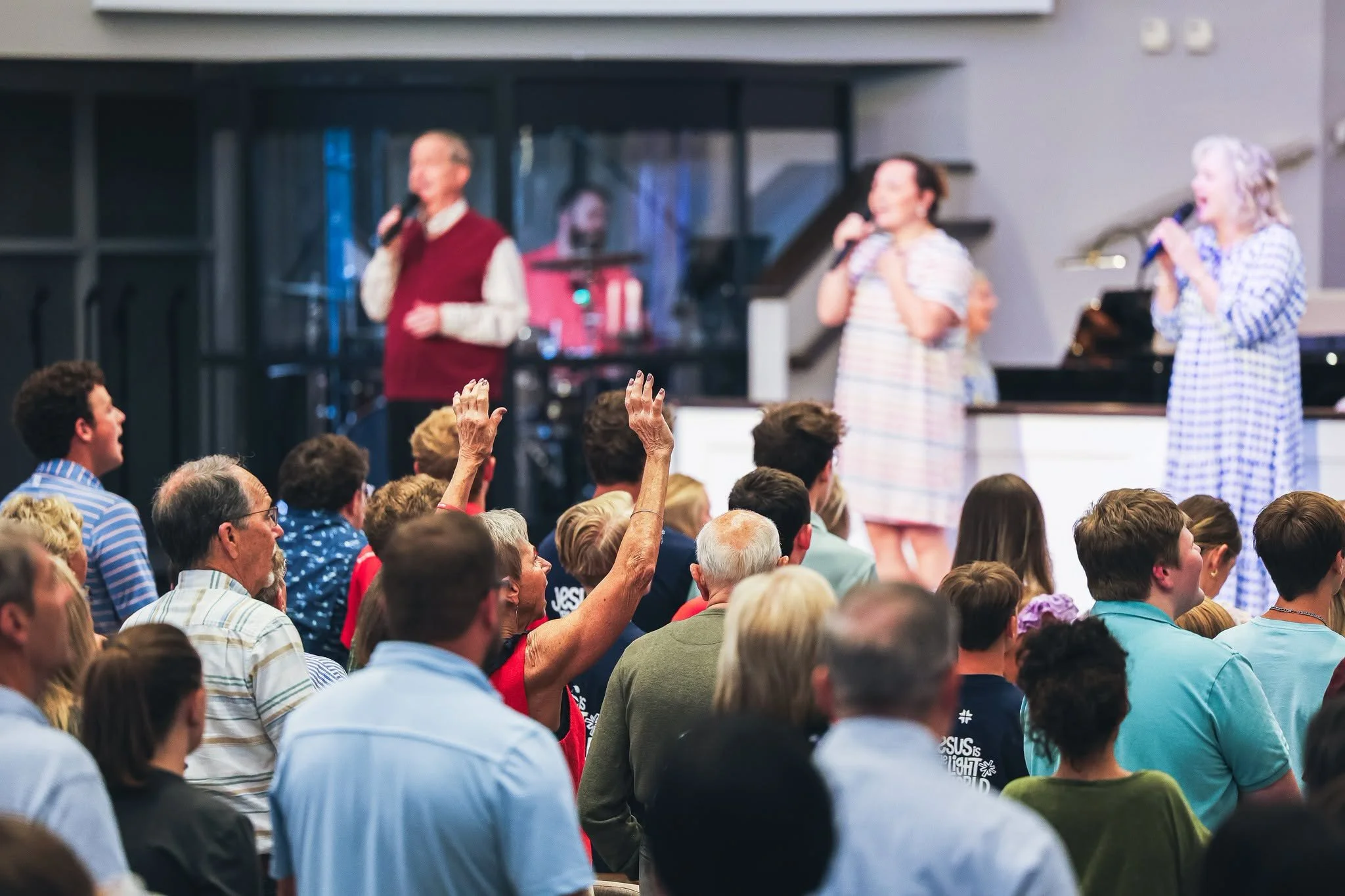 A large indoor gathering with many people seated and raising their hands. On stage, three women hold microphones and speak to the crowd, with a man playing keyboard in the background. The audience appears engaged, with some smiling and gesturing.