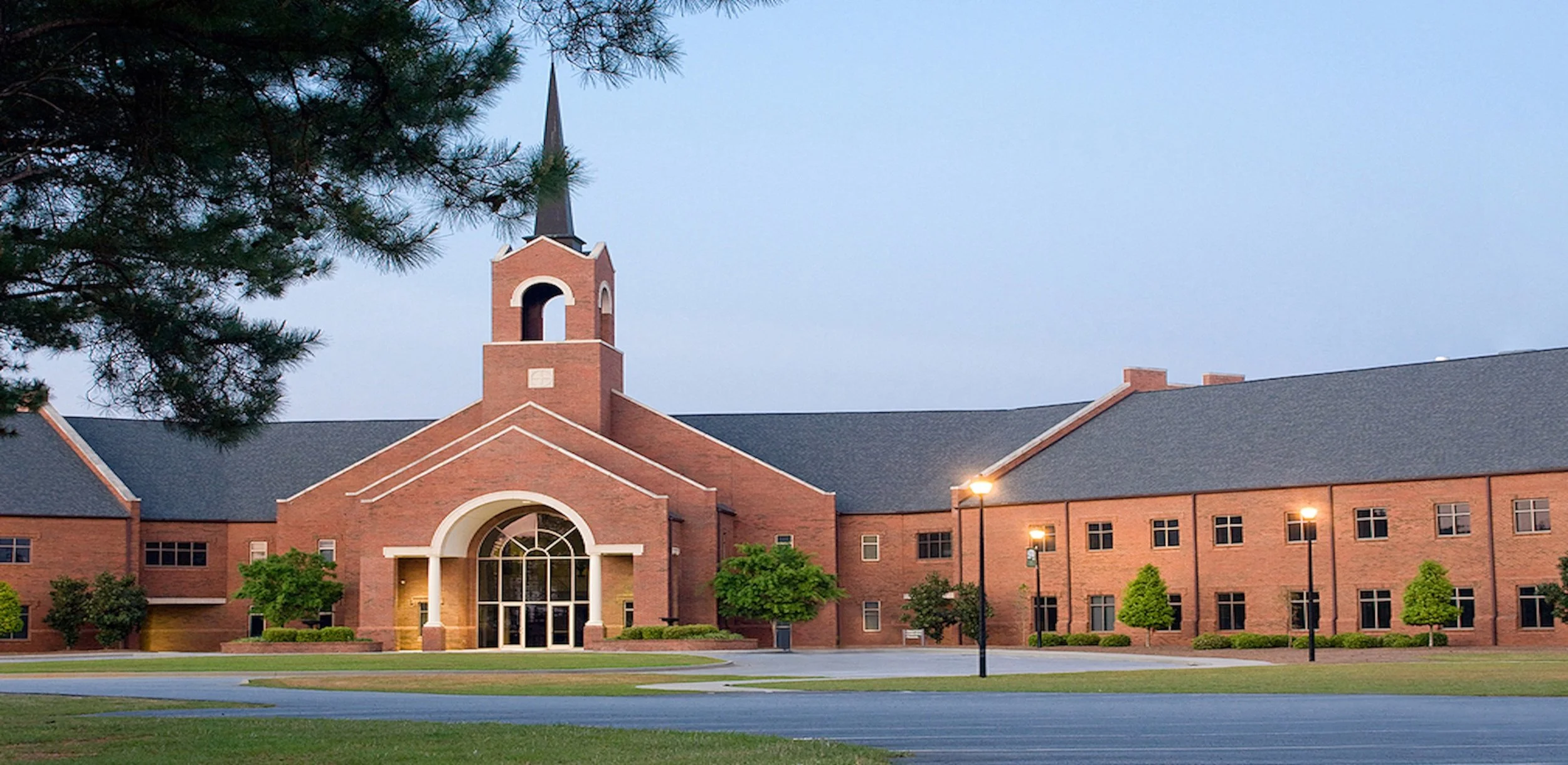 A large brick building with a central clock tower and a steeple, surrounded by green trees and lit street lamps, under a clear sky at dusk.