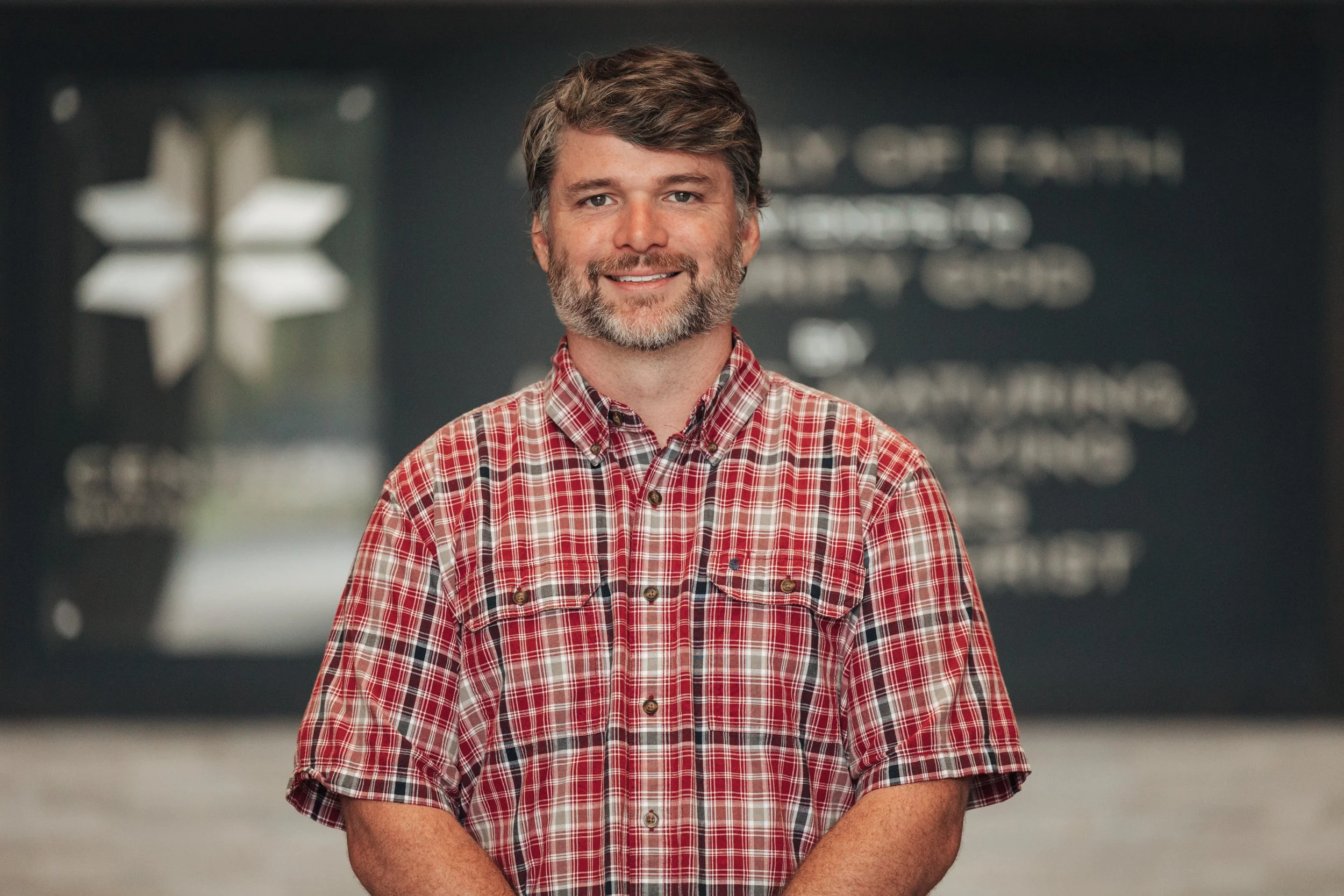 Portrait of a middle-aged man with a beard and brown hair, smiling, wearing a red and white plaid shirt, standing indoors with blurred background.