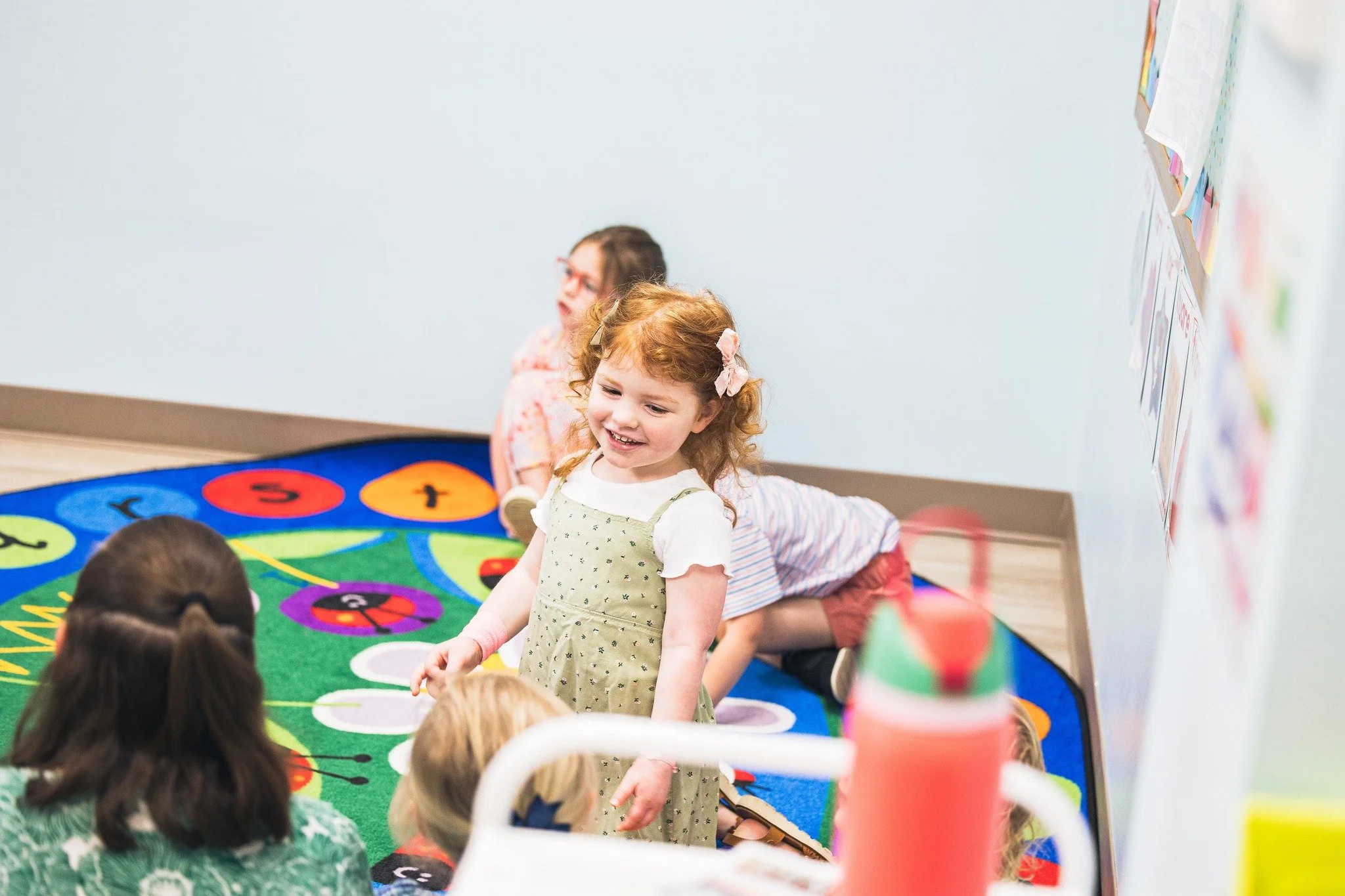Young girl with curly red hair and pink hair clips smiling and standing in a classroom with other children, colorful educational materials on the wall, and a colorful round rug on the floor.