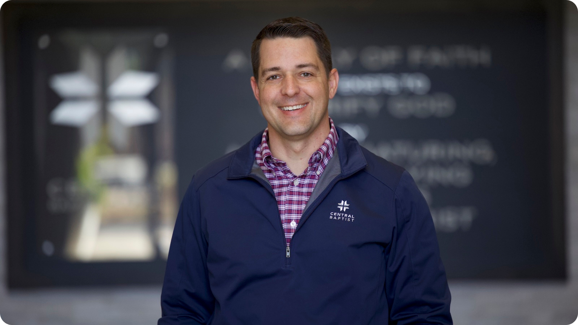 A man smiling in front of a blurred background with a church logo and text, wearing a navy blue jacket with a 'Central Baptist' logo and a red plaid shirt.