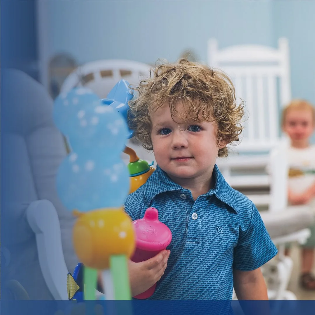 Young boy with curly hair wearing a blue shirt, holding a pink sippy cup, looking at the camera with a neutral expression, in a playroom or daycare setting with a blurred background of another child and nursery furniture.