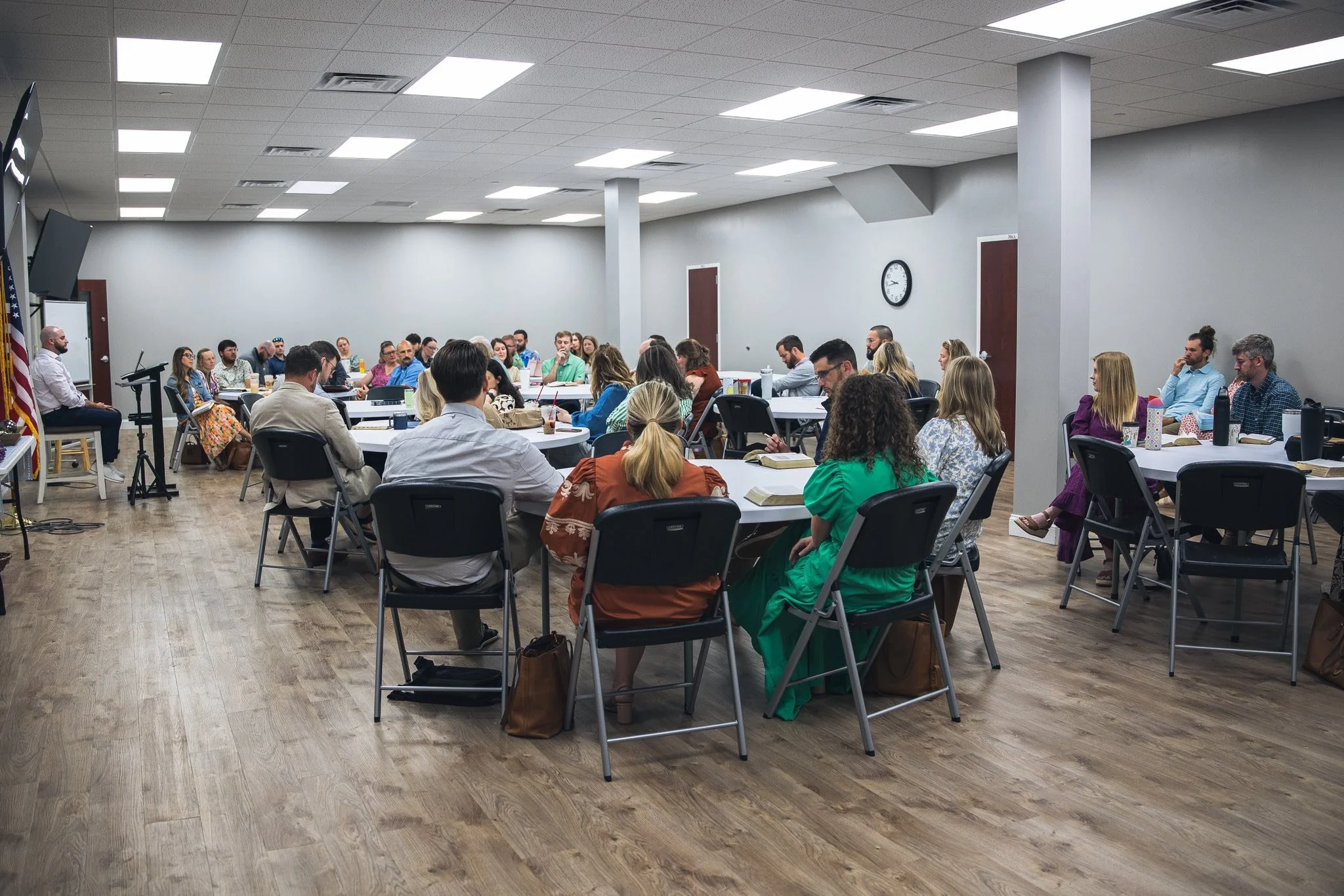 A group of people seated at round tables during a meeting or class in a conference room with a gray wall, a clock, and fluorescent lights.