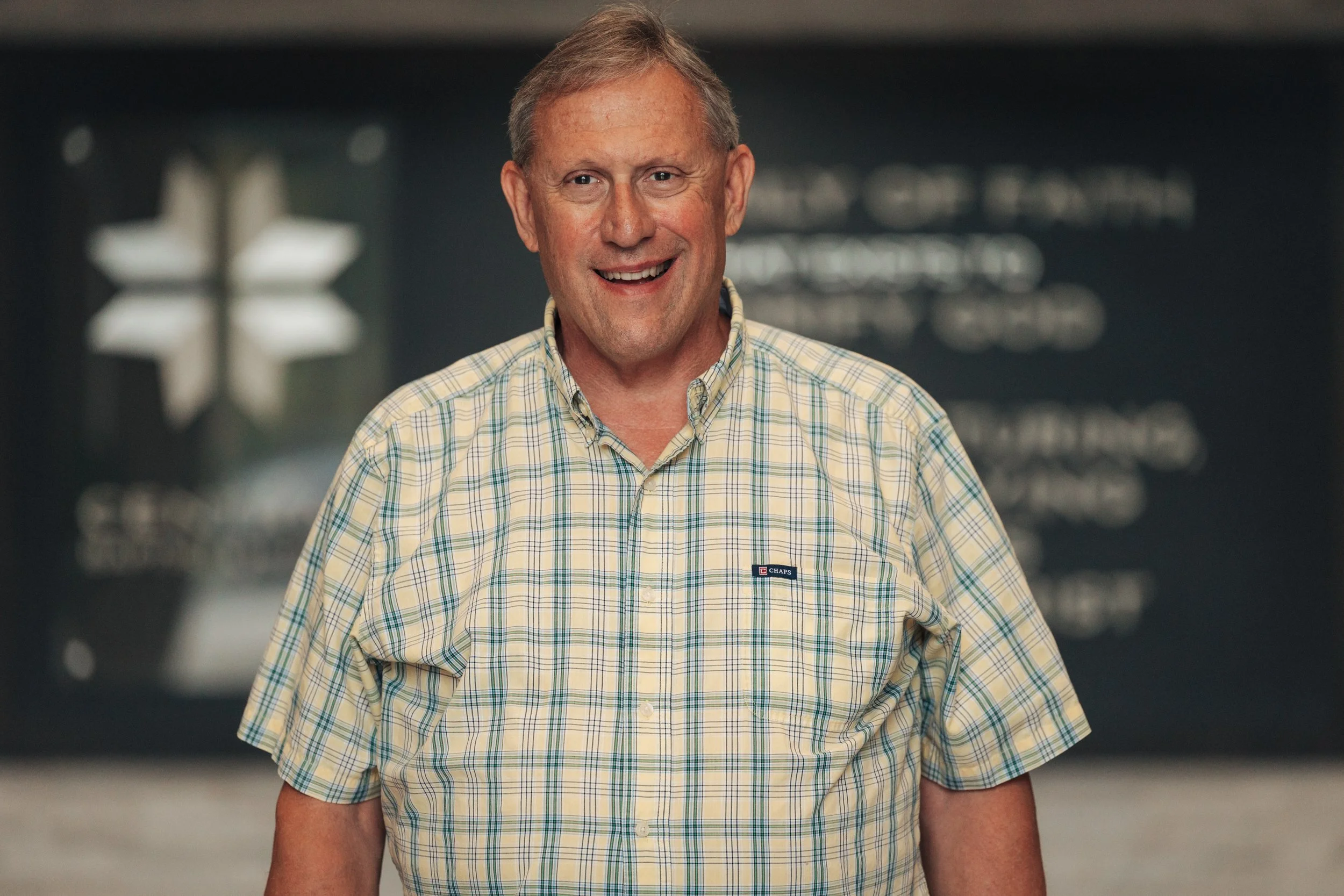 A middle-aged man with short, light-colored hair wearing a plaid shirt smiles at the camera. The background is blurred with indistinct text and symbols.