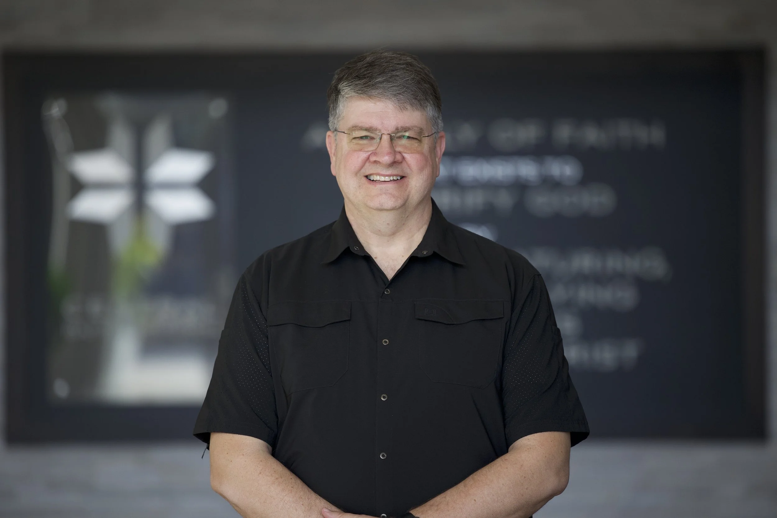 Portrait of a smiling man with glasses, wearing a black shirt, standing in front of a blurred background with text and geometric shapes.
