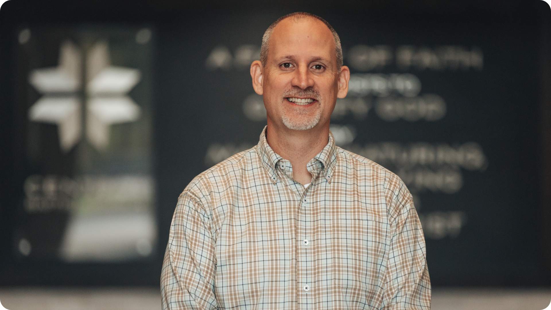 A smiling man with a buzz cut wearing a beige checkered shirt stands in front of a blurred background with indistinct text and a logo.