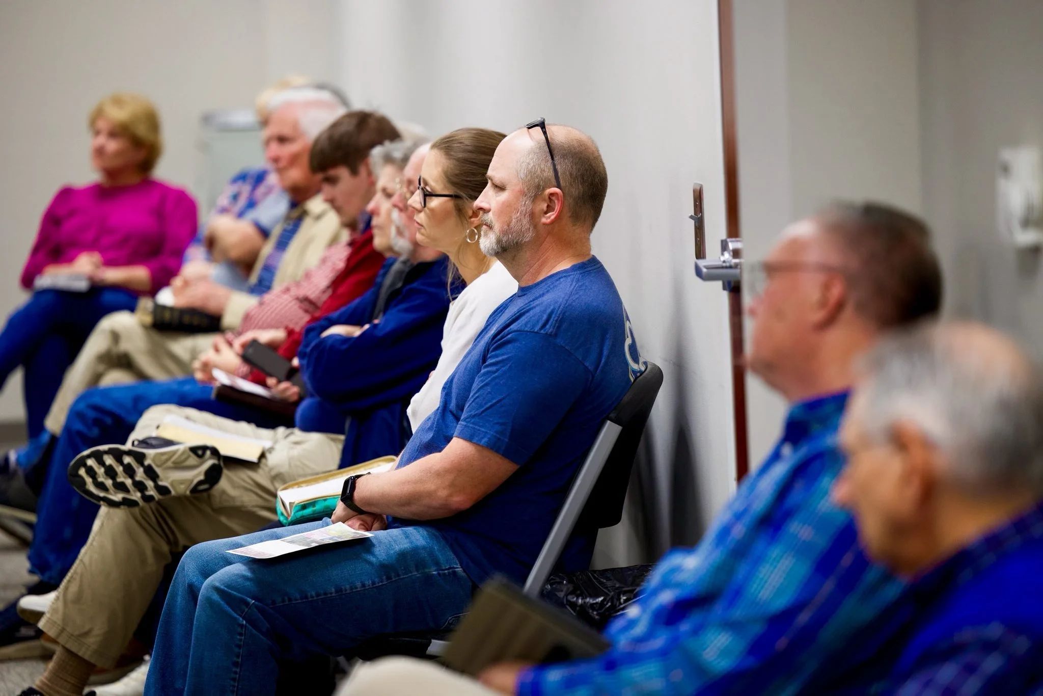 A group of people sitting in a row at an indoor event, focused and listening. The background shows a plain wall.