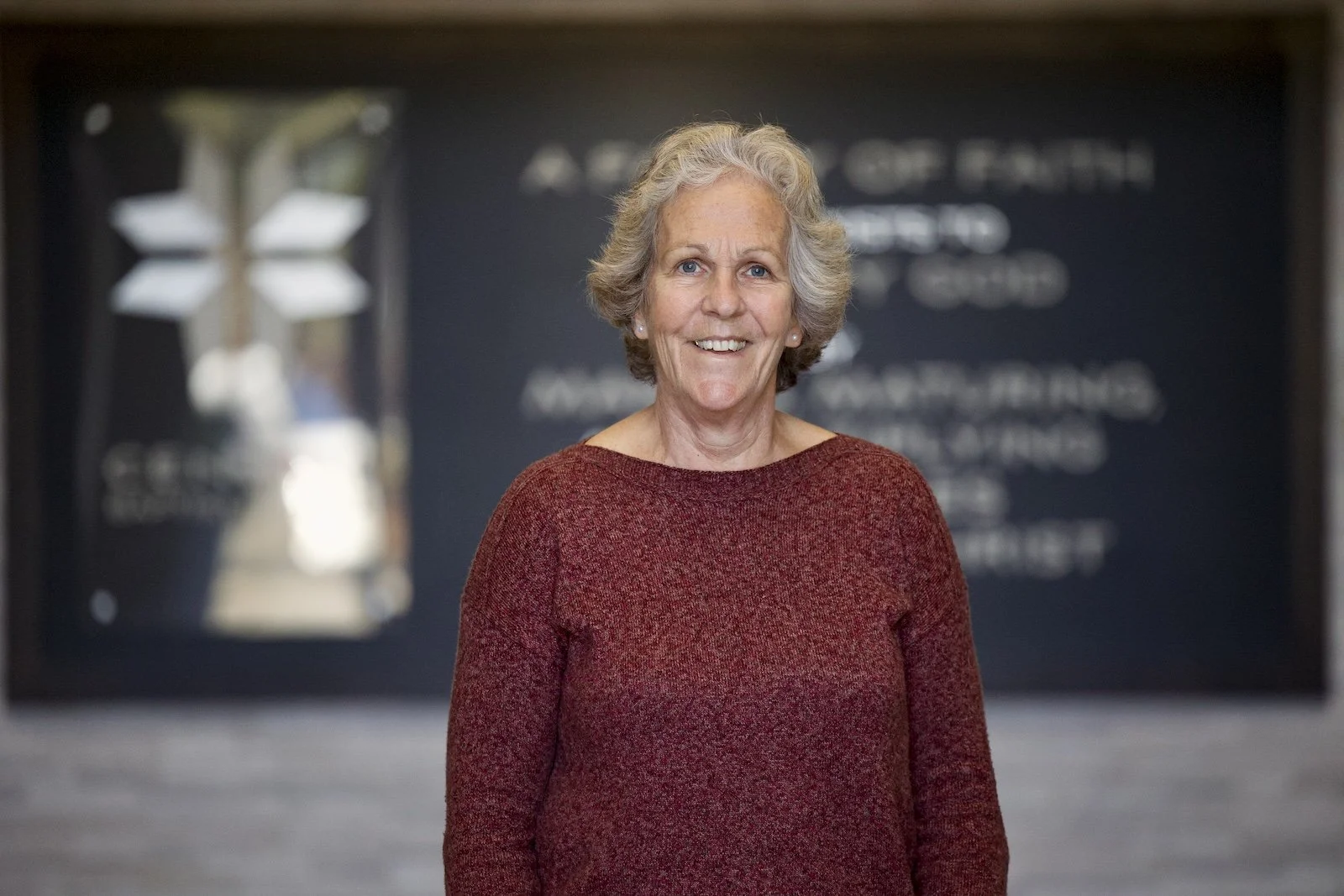 Smiling elderly woman with short gray hair wearing a red sweater, standing indoors against a dark background with out-of-focus text and a decorative element.