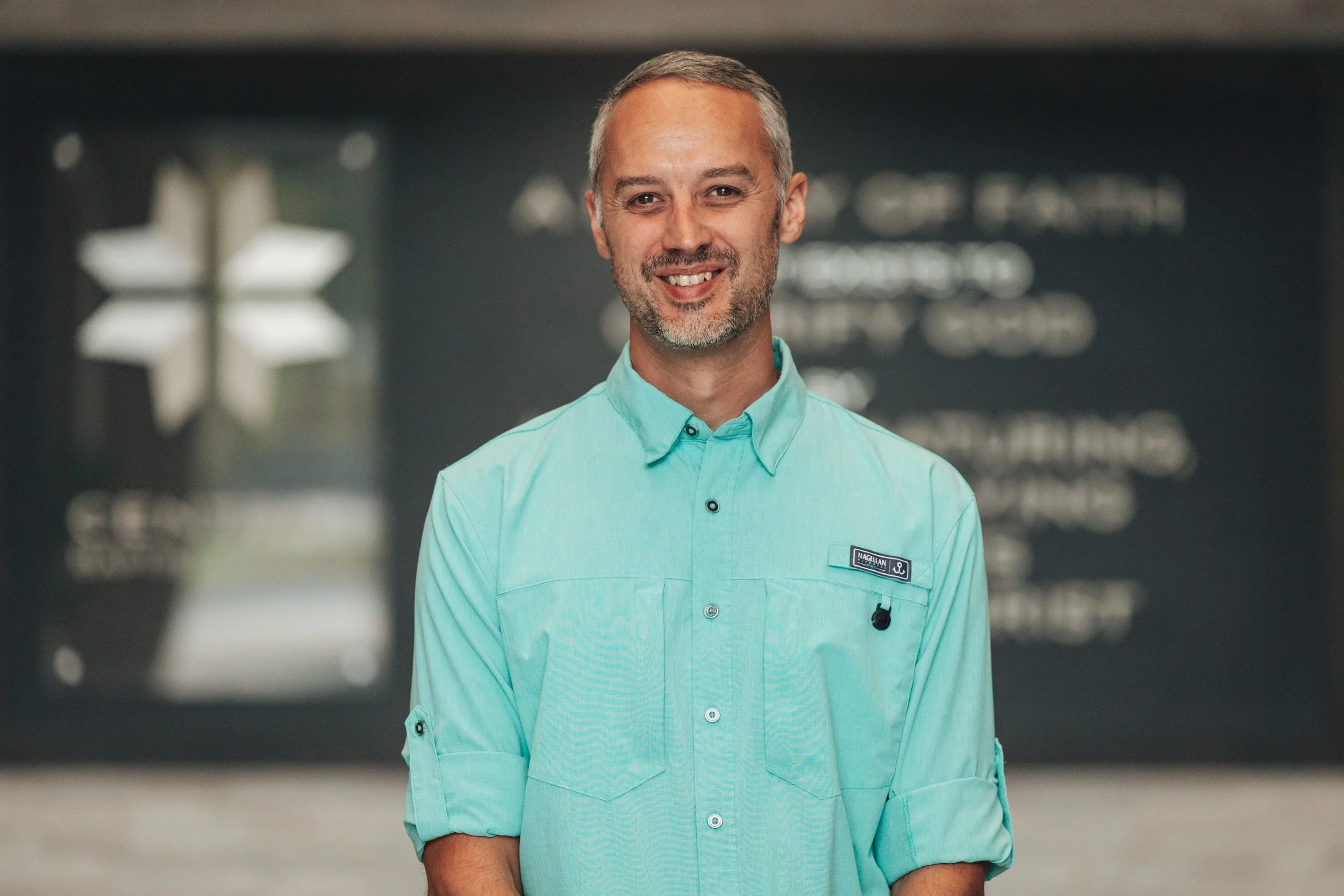 A smiling middle-aged man with short gray hair and a beard, wearing a turquoise uniform shirt, standing in an indoor setting with a blurred background.