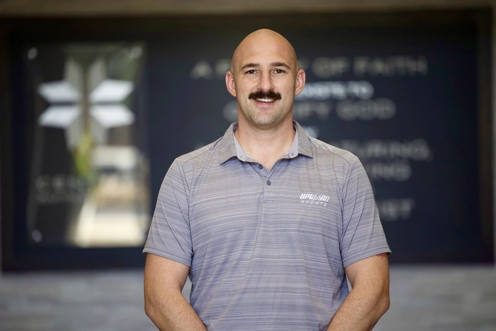 A man with a bald head and mustache standing indoors, wearing a gray golf shirt with an UPHD Sports logo on it, smiling at the camera. The background features a blurred blackboard and some décor.