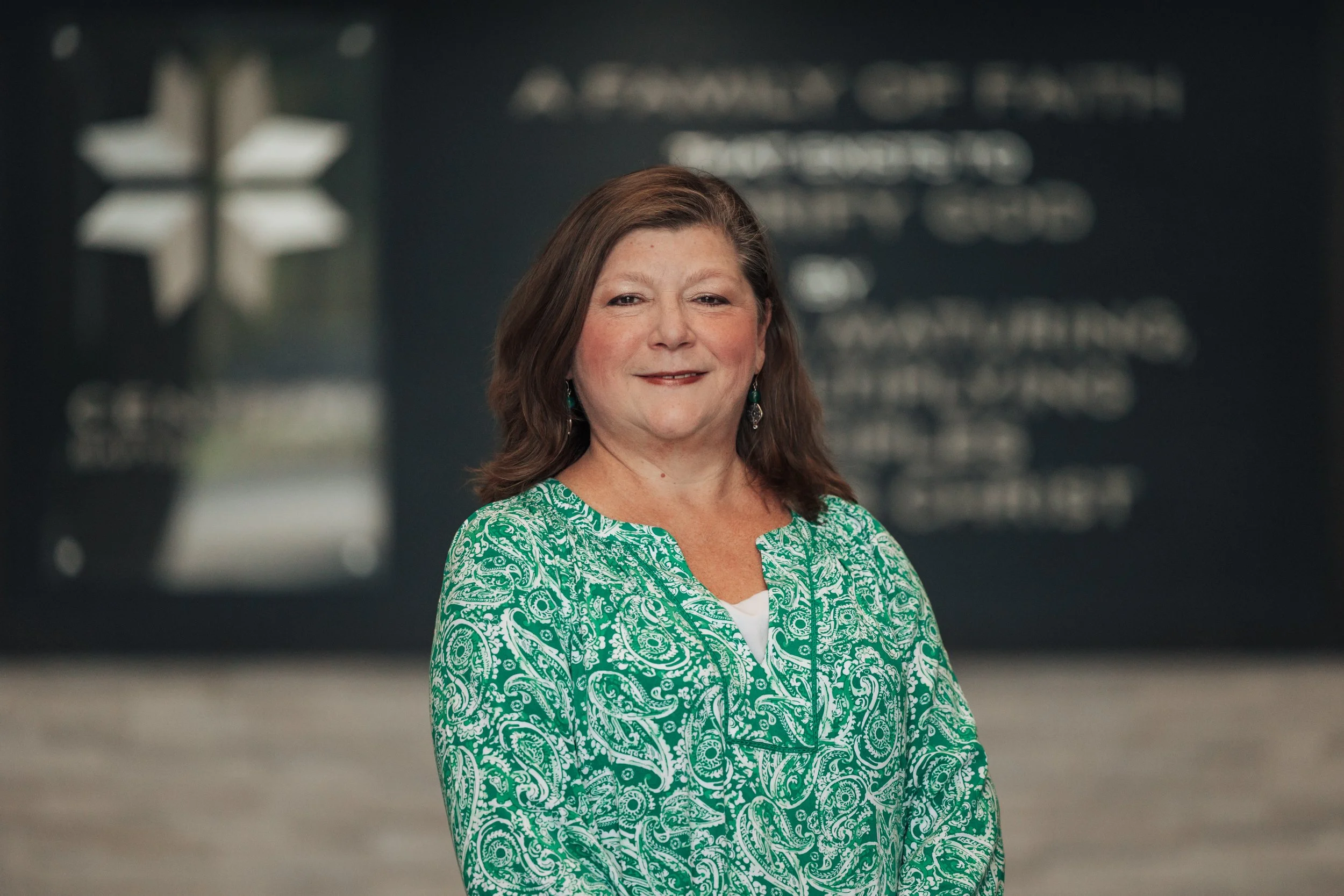 A woman with shoulder-length brown hair wearing a green paisley-patterned blouse and earrings, standing in an indoor setting with blurred background and signage.