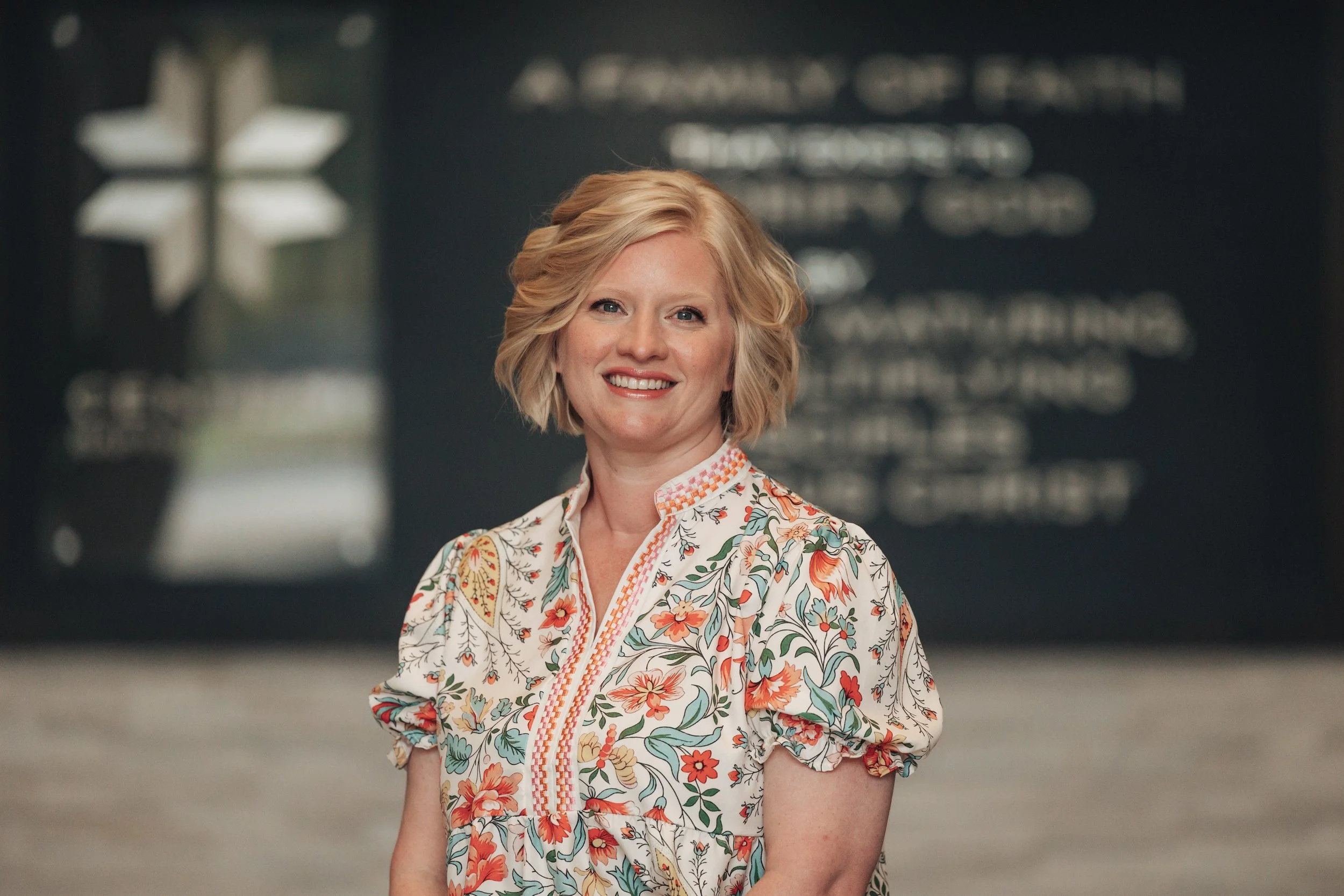 A woman with blonde hair smiling in a colorful floral shirt standing in an indoor setting with a dark blurred background.