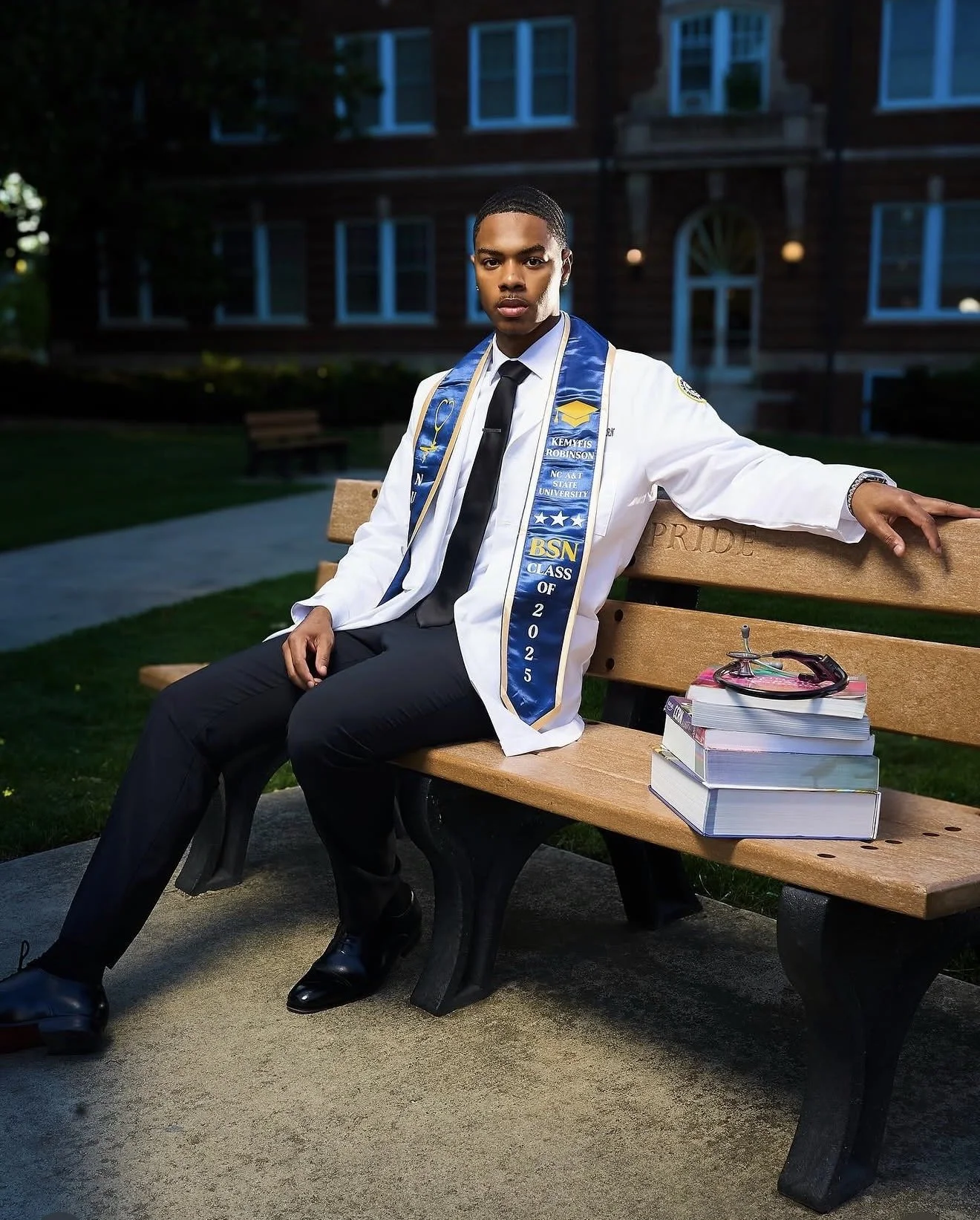 Young man in graduation cap and gown sitting on a bench outside at night, with a stack of books and a stethoscope next to him.