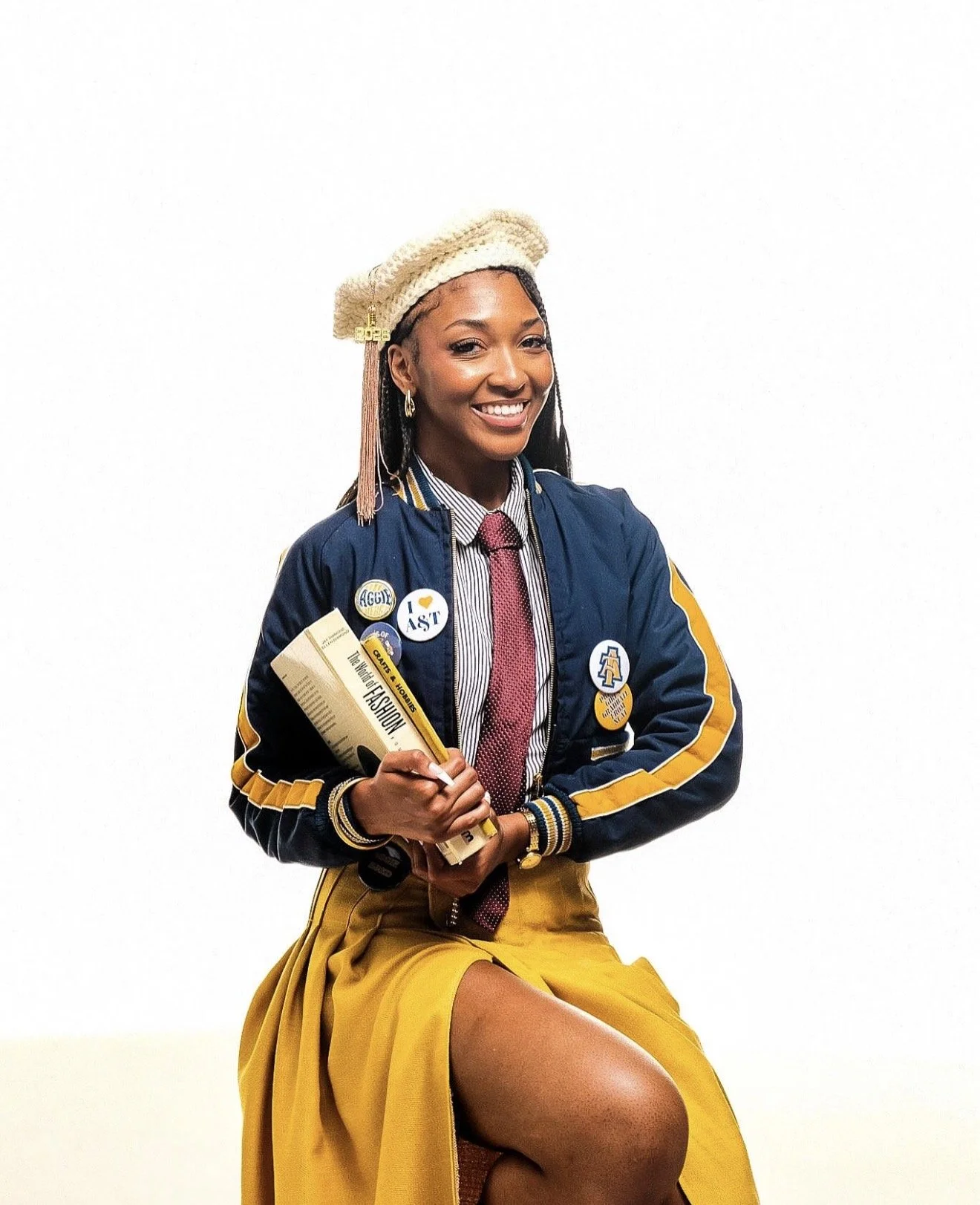 Young woman smiling, wearing a blue and yellow letterman jacket with pins, a striped shirt, a pink tie, a cream-colored beret, and sitting against a plain white background, holding a book titled "The Little Black Fashion Book."
