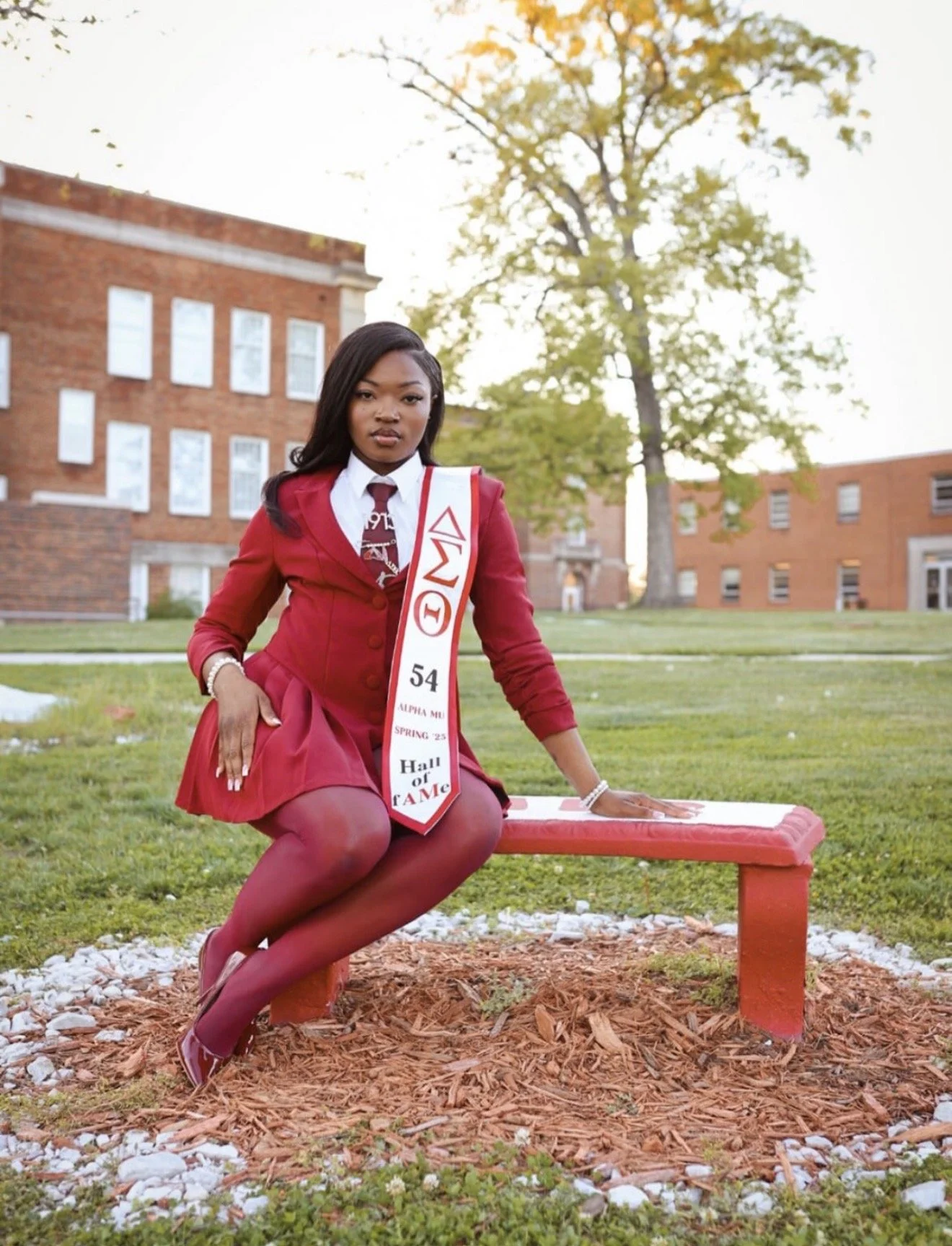 A young woman dressed in a red junior sorority jacket, skirt, and tights, sitting on a red park bench outside on a grassy lawn with a university campus in the background. She is wearing a white shirt, red tie, and a sorority sash that reads 'Alpha Phi,' with a title 'Hall of Fame' and the number '54'. There is a large tree behind her and brick buildings in the background.