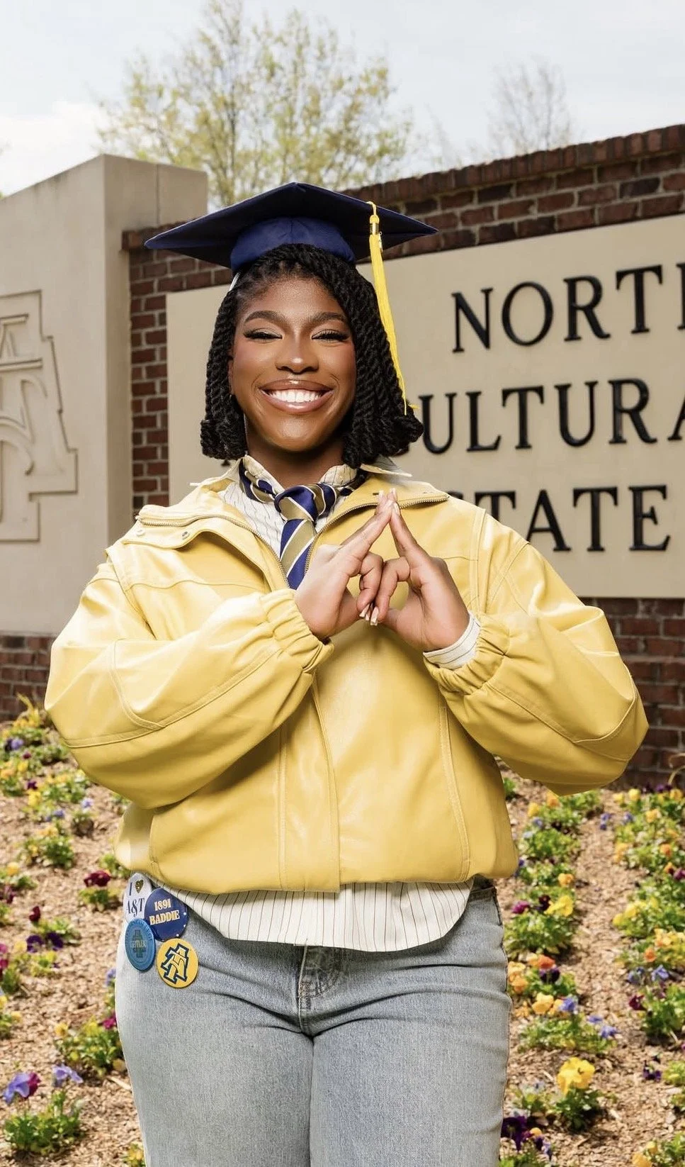 Young woman in yellow graduation gown and cap, smiling with hands in prayer gesture, standing outside North Carolina State University sign and flowering garden.