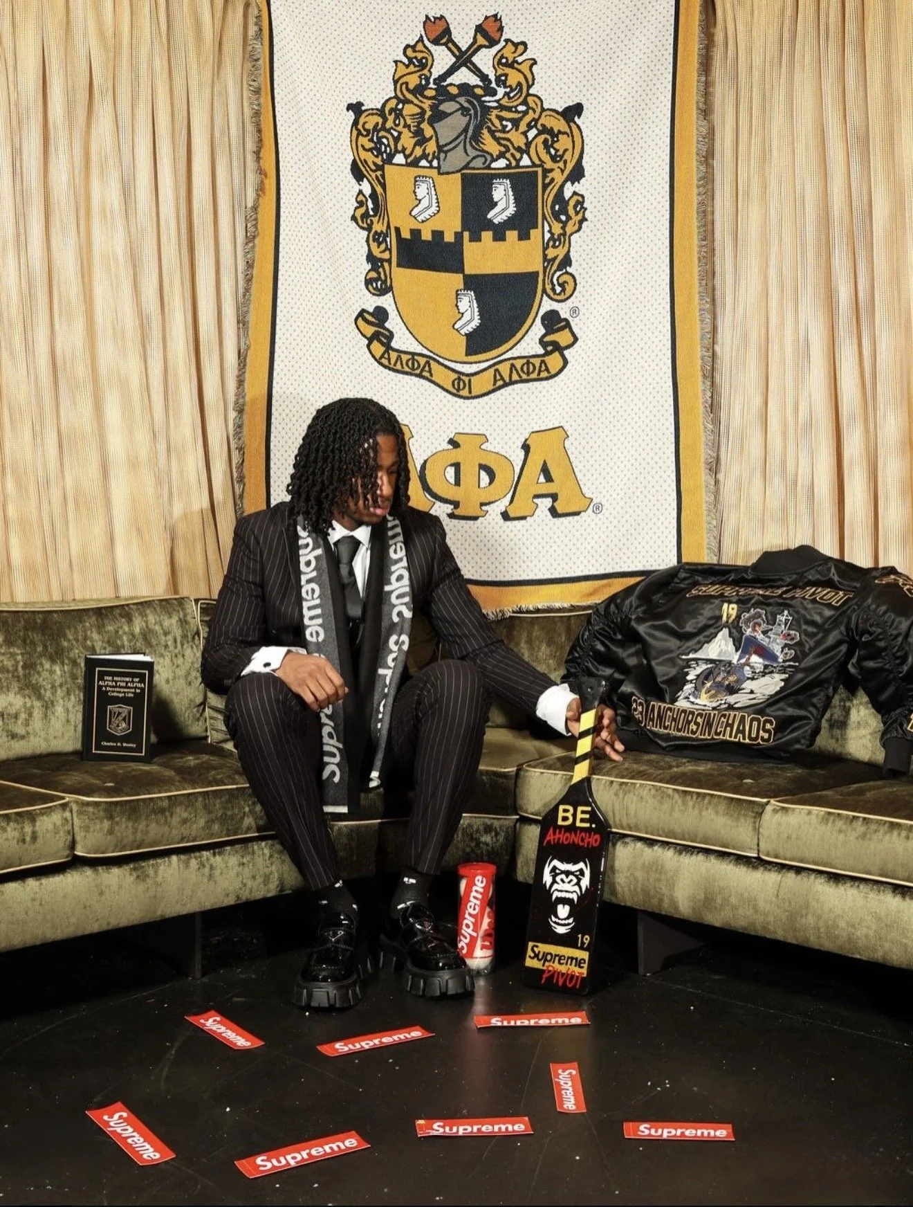 A person dressed in a pinstripe suit with black platform shoes is seated on a vintage green velvet couch, holding a hockey stick. The scene features a backdrop with the Alpha Phi Alpha crest, and the individual is surrounded by Supreme-branded items,