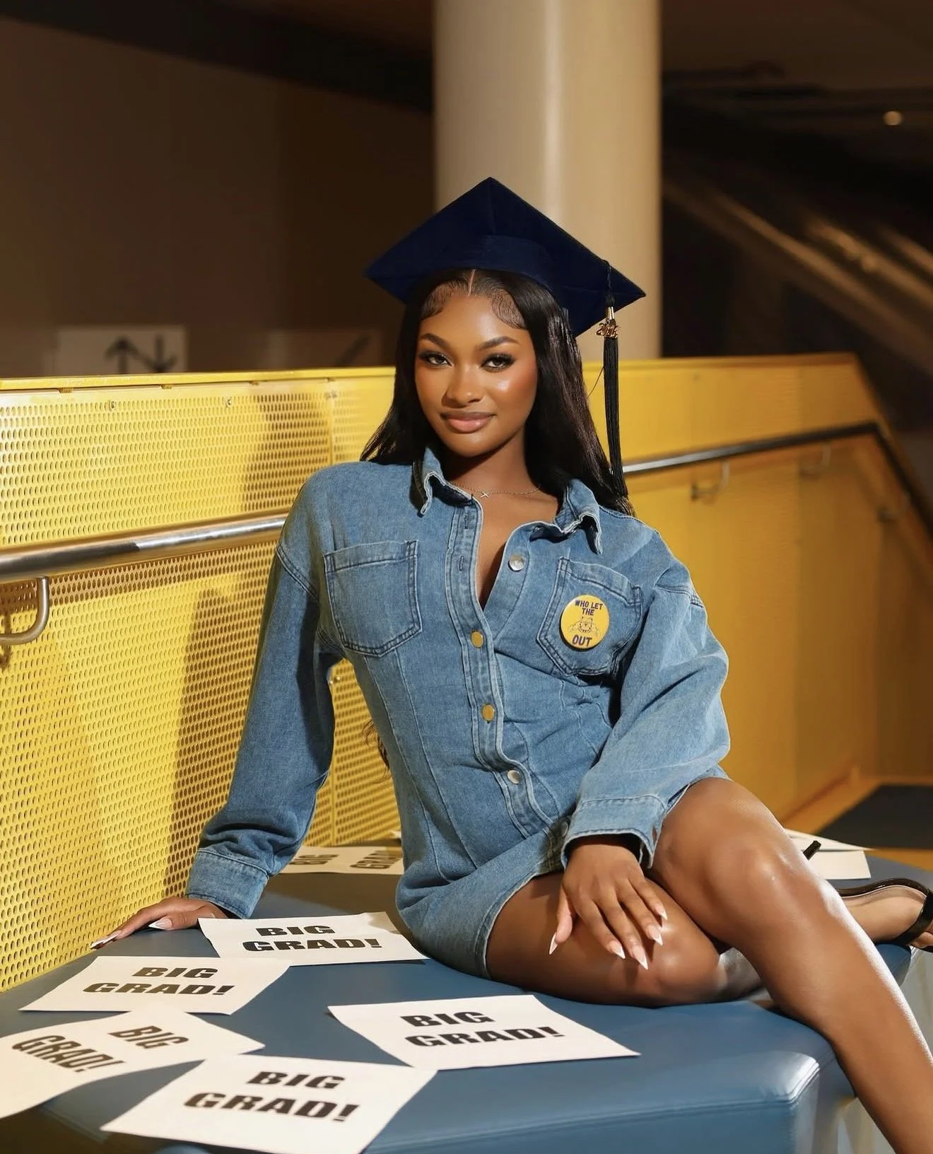 A young woman in a denim jacket and graduation cap sitting on a table with "Big Grad!" signs in front of her in a modern indoor setting.
