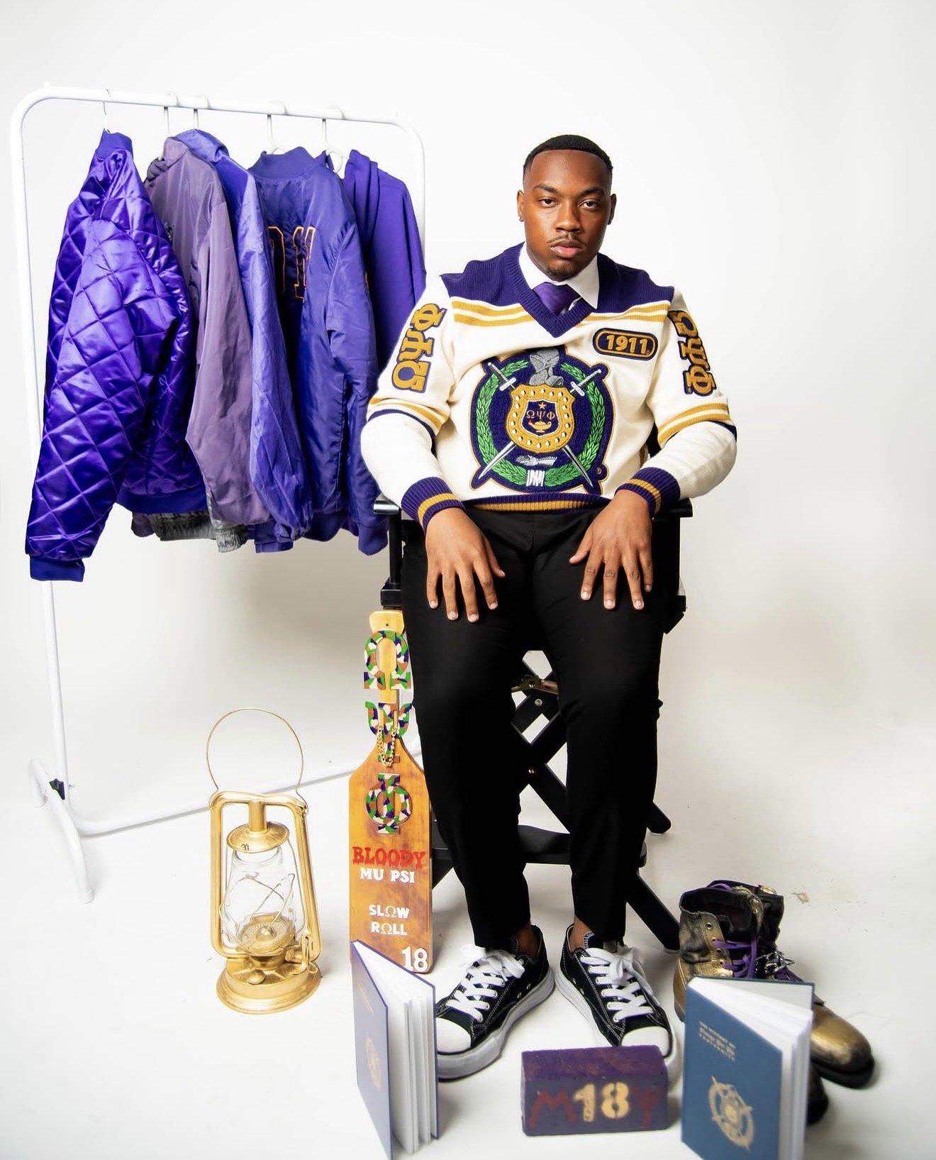 A young man sitting on a black chair in front of a white background with a clothing rack of purple jackets behind him. On the floor in front of him are various objects, including books, a gold clock, a colorful skateboard, a water bottle, and shoes. 