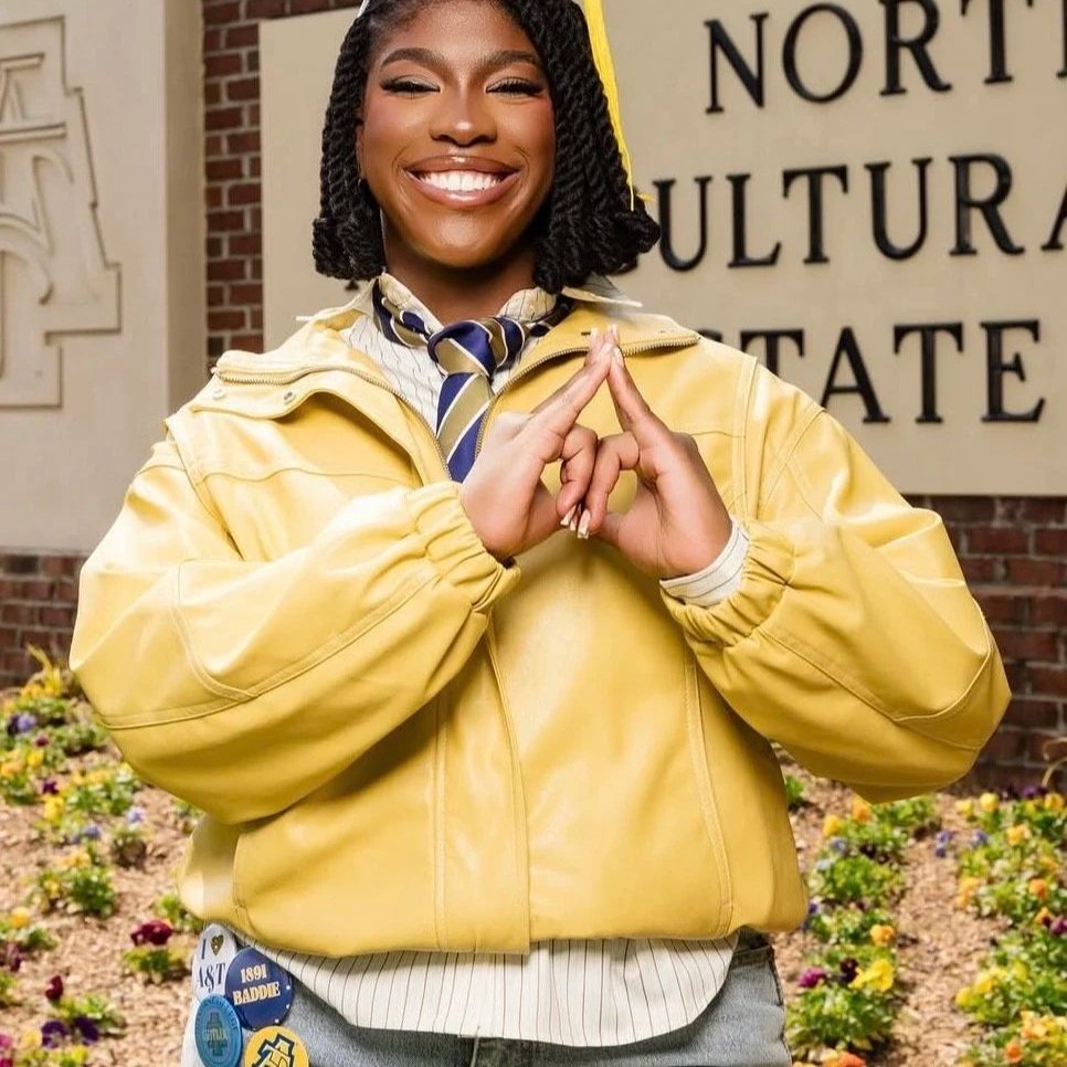 A young woman with braids smiling and making a gesture with her hands, wearing a yellow jacket and standing in front of a sign that says 'North Cultural State' with flowers in the foreground.
