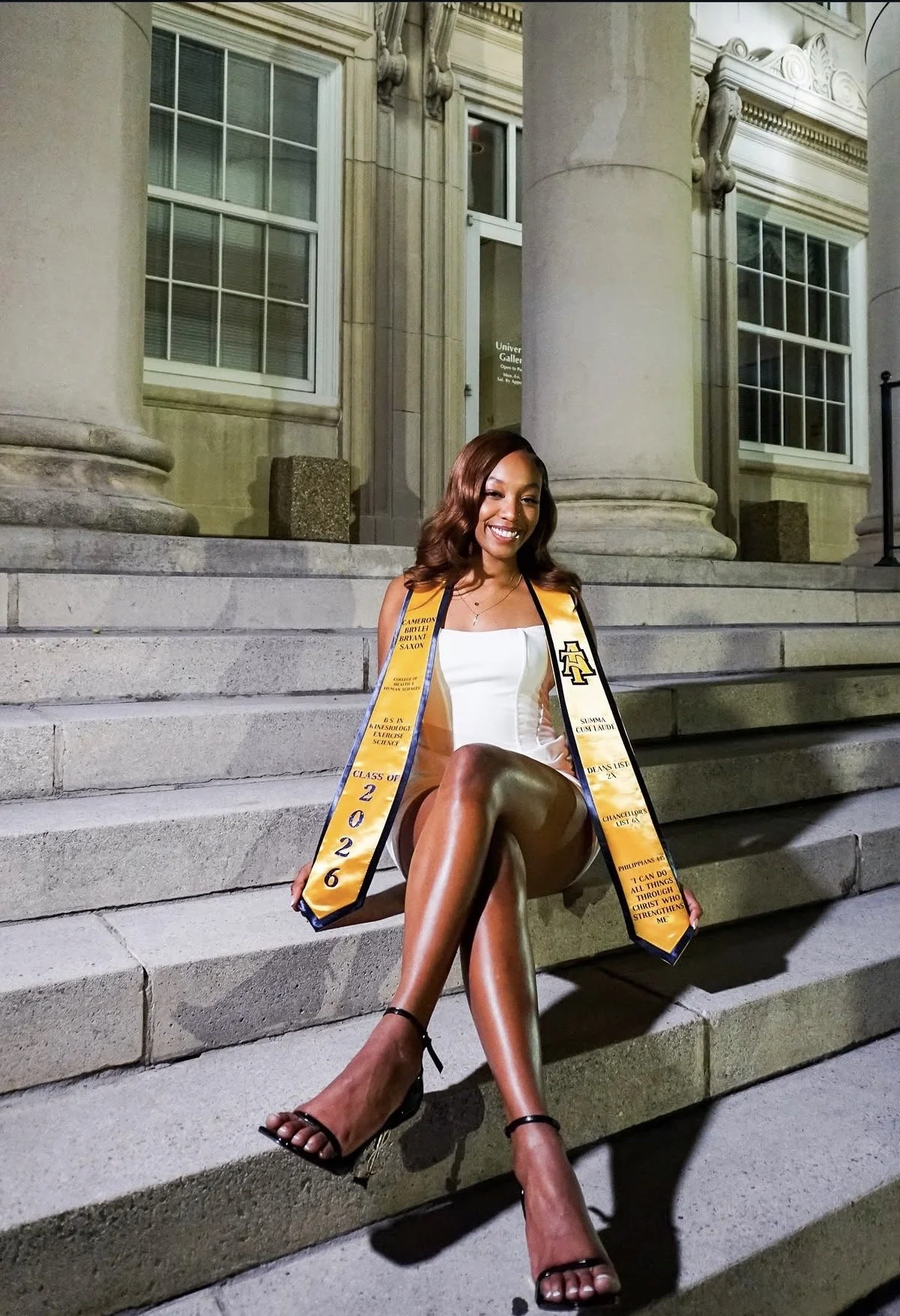 A young woman with long, wavy brown hair smiles while sitting on the steps of a historic building with large columns. She is wearing a white dress and black high heels, and a yellow and black academic stole that reads 'Class of 2026' and other gradua