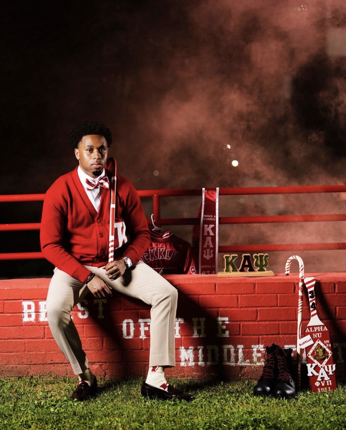 A young man dressed in a red cardigan, white shirt, beige pants, and a red and white bow tie is sitting on a red brick wall. He holds a red and white cane. Surrounding him are Greek letters and sorority paraphernalia, including a pair of black shoes,