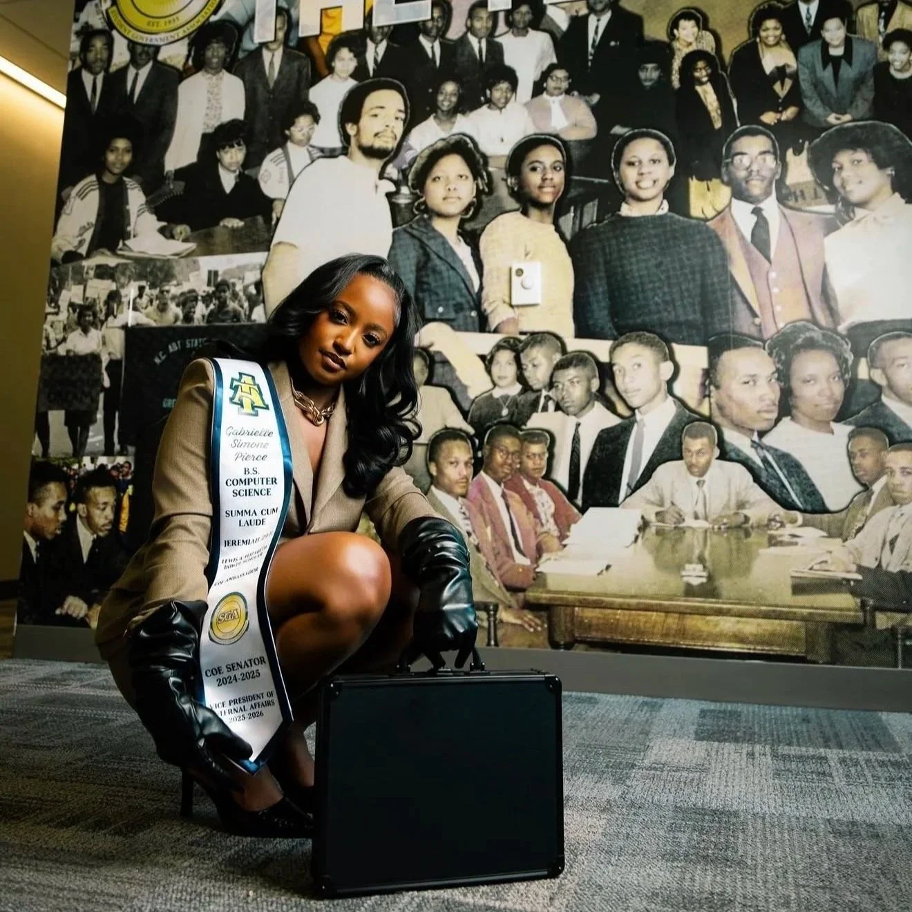 A woman wearing a sash and black gloves kneeling in front of a large collage photo, which includes a mix of black and white and color images of people in a formal setting. The woman has long black hair and is dressed in a beige outfit with high heels, and she is squatting next to a black briefcase on the ground.
