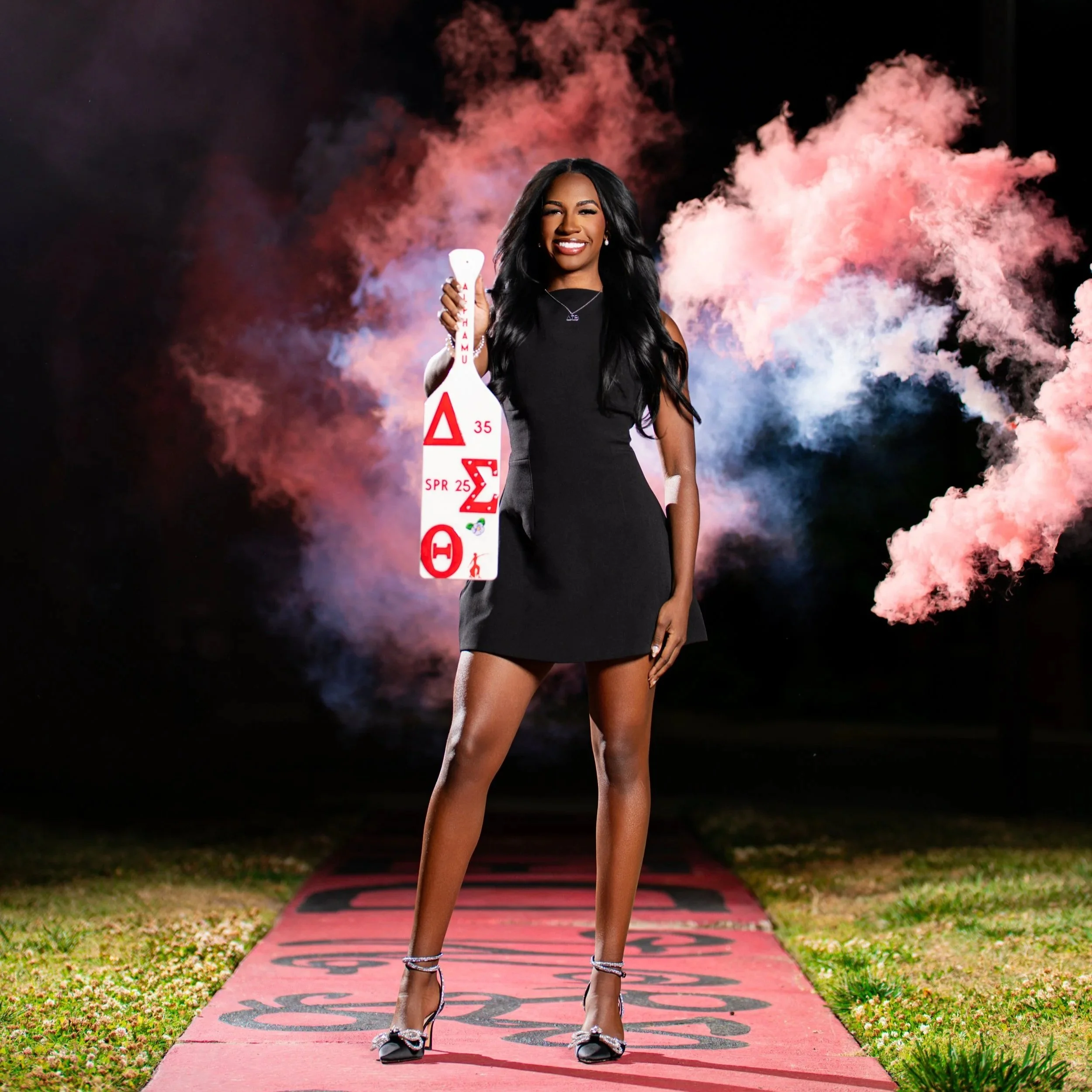 Young woman smiling and posing on a pink sidewalk with black numbers, holding a white object with red and black markings. She is dressed in a black dress, wearing high heels, and standing against a backdrop of pink and purple smoke in the night sky.
