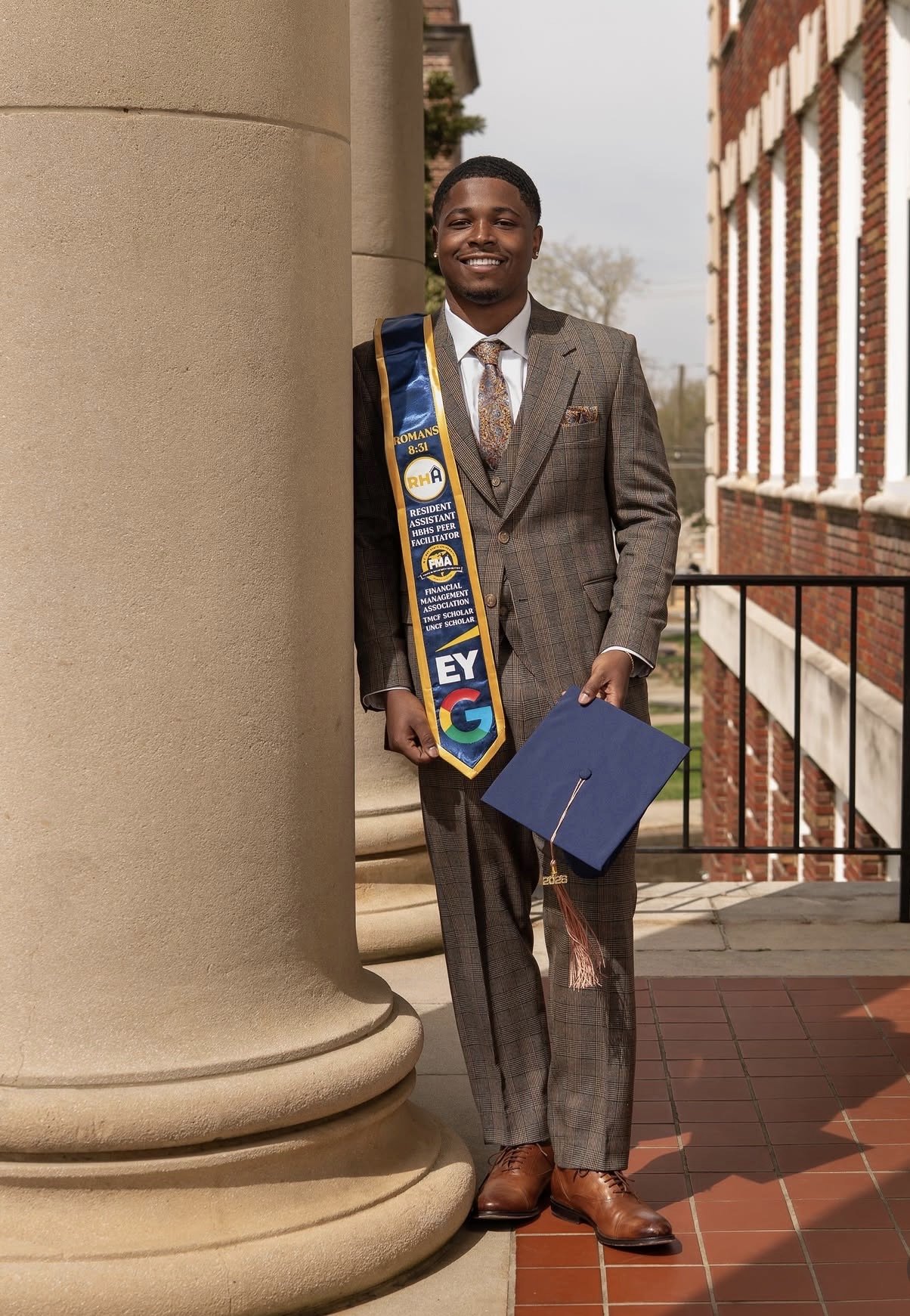 Smiling young man in a suit holding a blue graduation cap, standing outdoors near a large column and brick building.