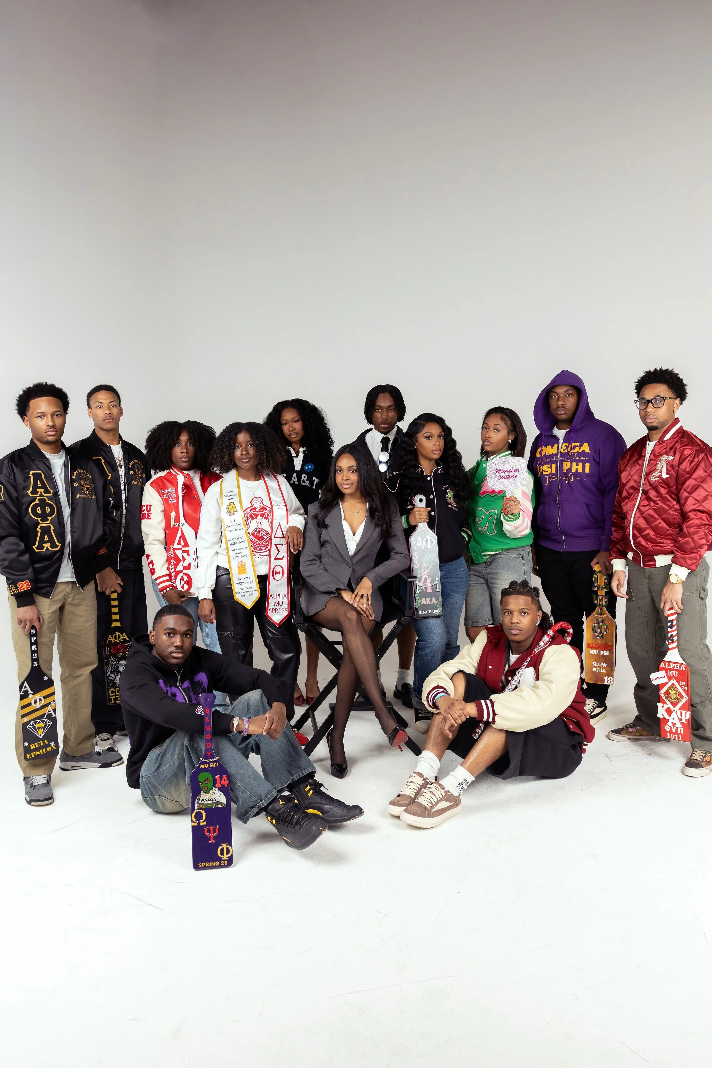 Group of diverse young adults posing in a photo studio, some sitting and some standing, wearing casual streetwear and sporting sorority paraphernalia, with a plain white background.