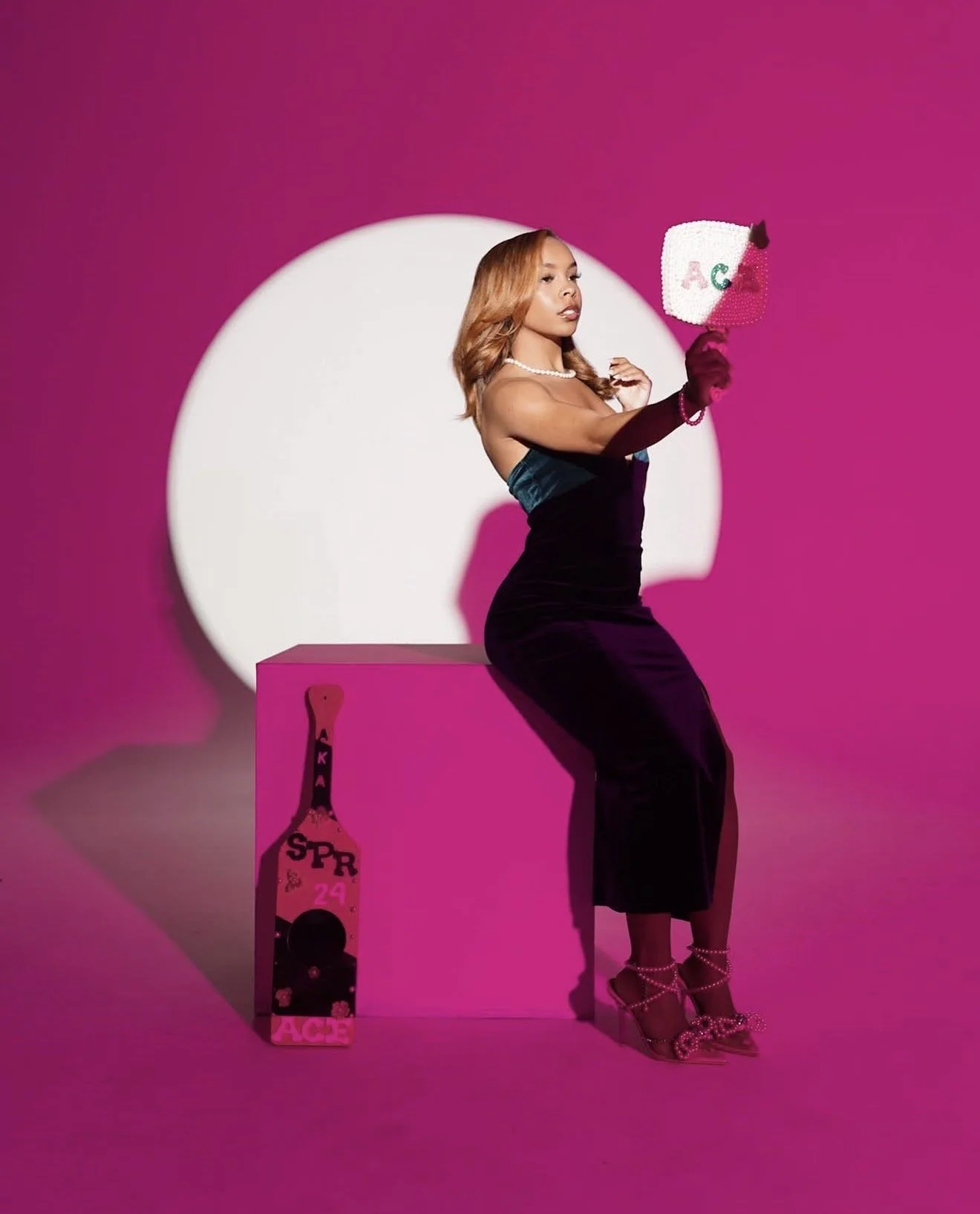 A woman with blonde hair in a black strapless dress sitting on a pink block, holding a sequined purse with green and red letters, surrounded by pink background and pink props including a box with a bottle illustration and the word 'ACE', and pink hig