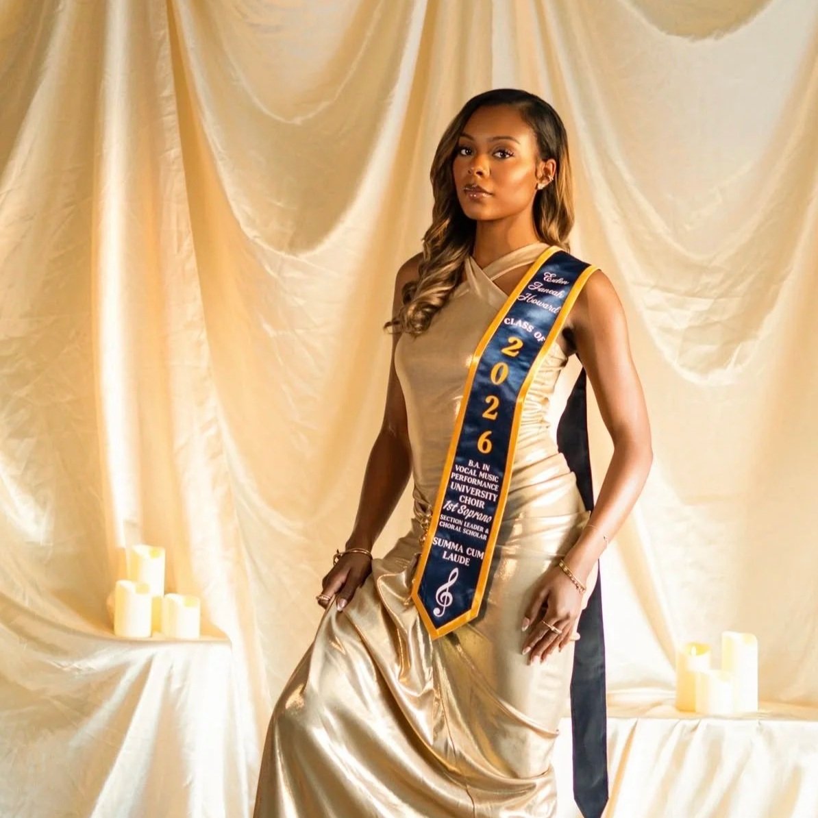 A young woman in a gold gown stands in front of a cream-colored draped backdrop, wearing a sash that reads 'Class of 2026' and features academic honors and achievements.