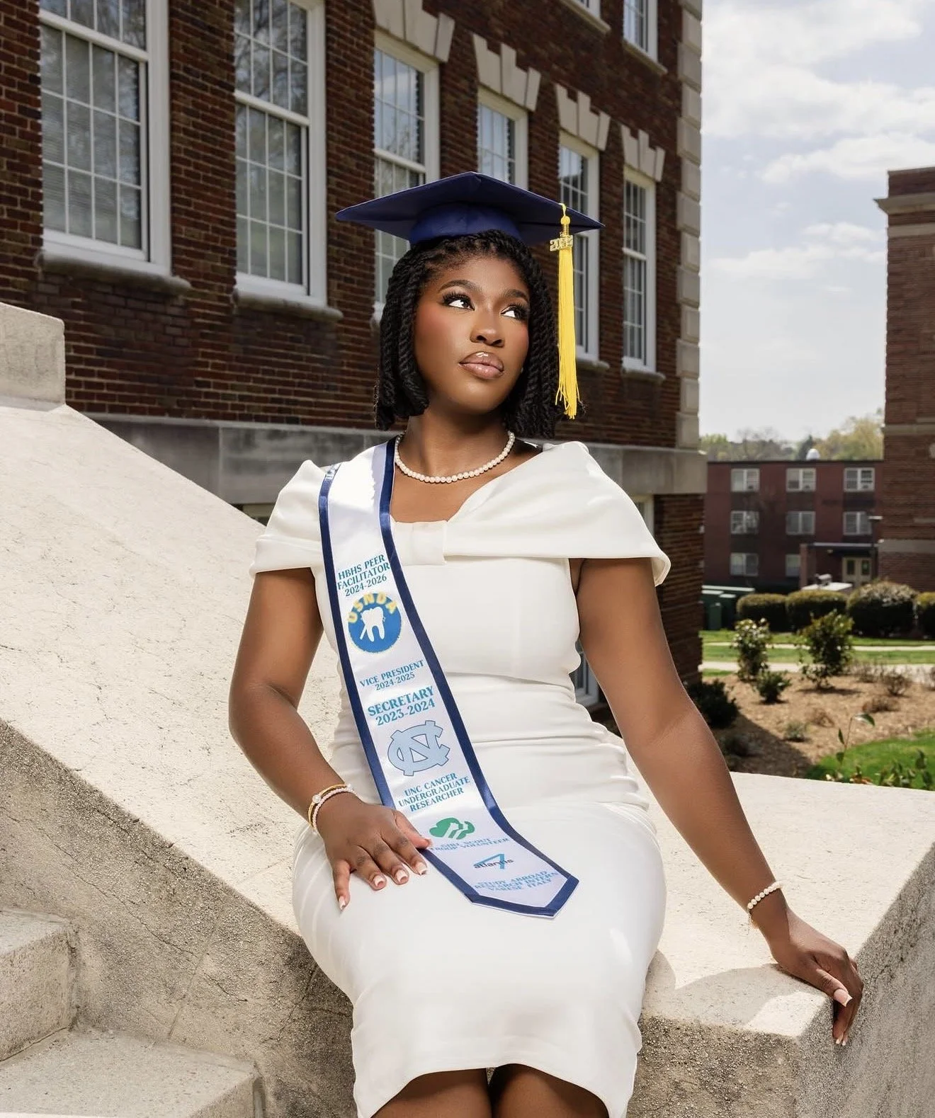 A young woman in a white dress wearing a blue graduation cap with a yellow tassel and a sash with various titles and logos, sitting on concrete stairs outside a brick building with trees and bushes in the background.