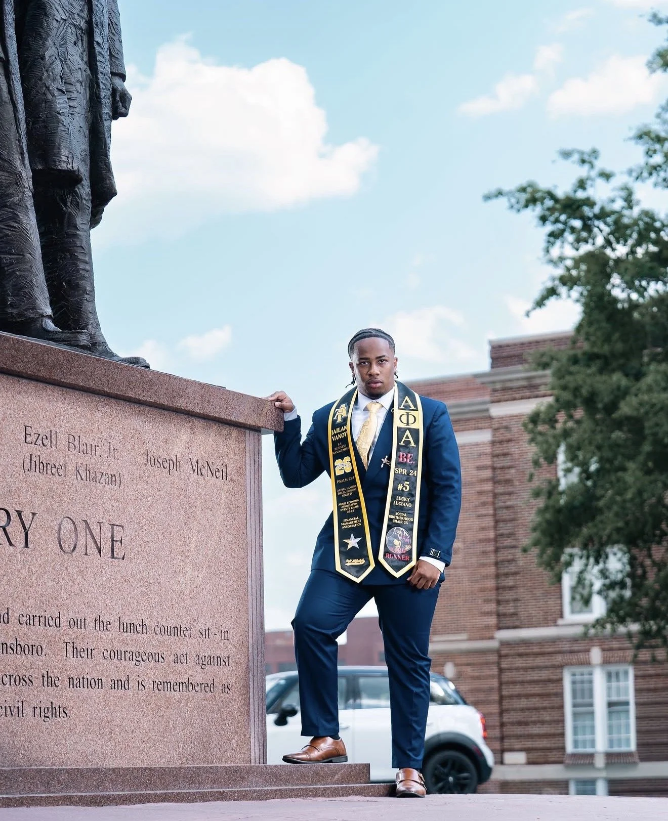A young man in a blue suit and tie standing beside a monument, wearing graduation cords and medals. He is looking at the camera with a serious expression, with greenery and brick buildings in the background.