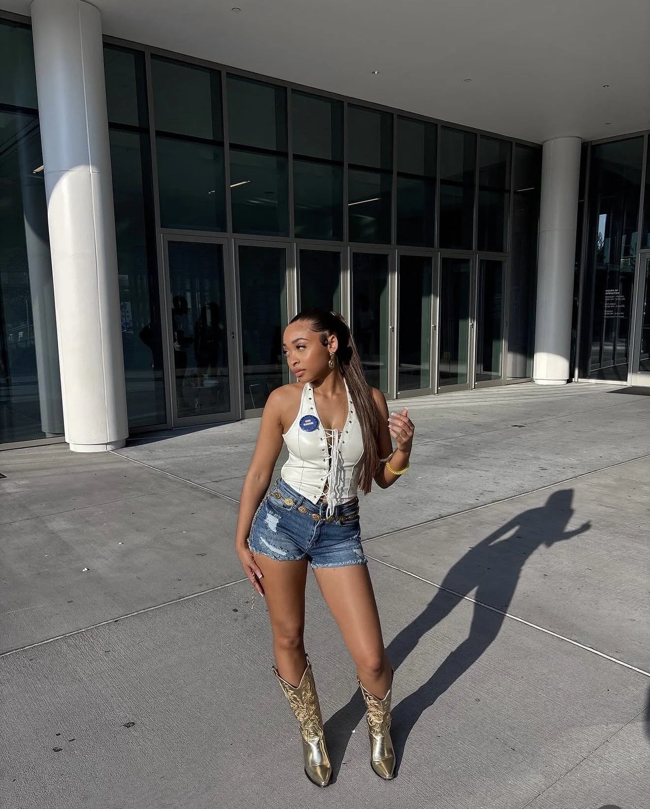 A woman standing outside a modern building with large glass windows, wearing a white lace-up top, distressed denim shorts, and gold cowboy boots, holding a phone, with sunlight casting shadows.