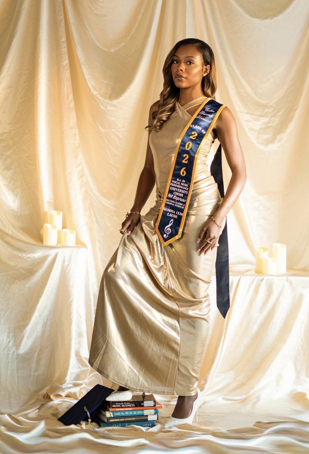 A woman in a gold evening gown standing in front of gold drapes with lit candles on a table. She wears a sash that reads 'Class of 2022' and holds books on music business.