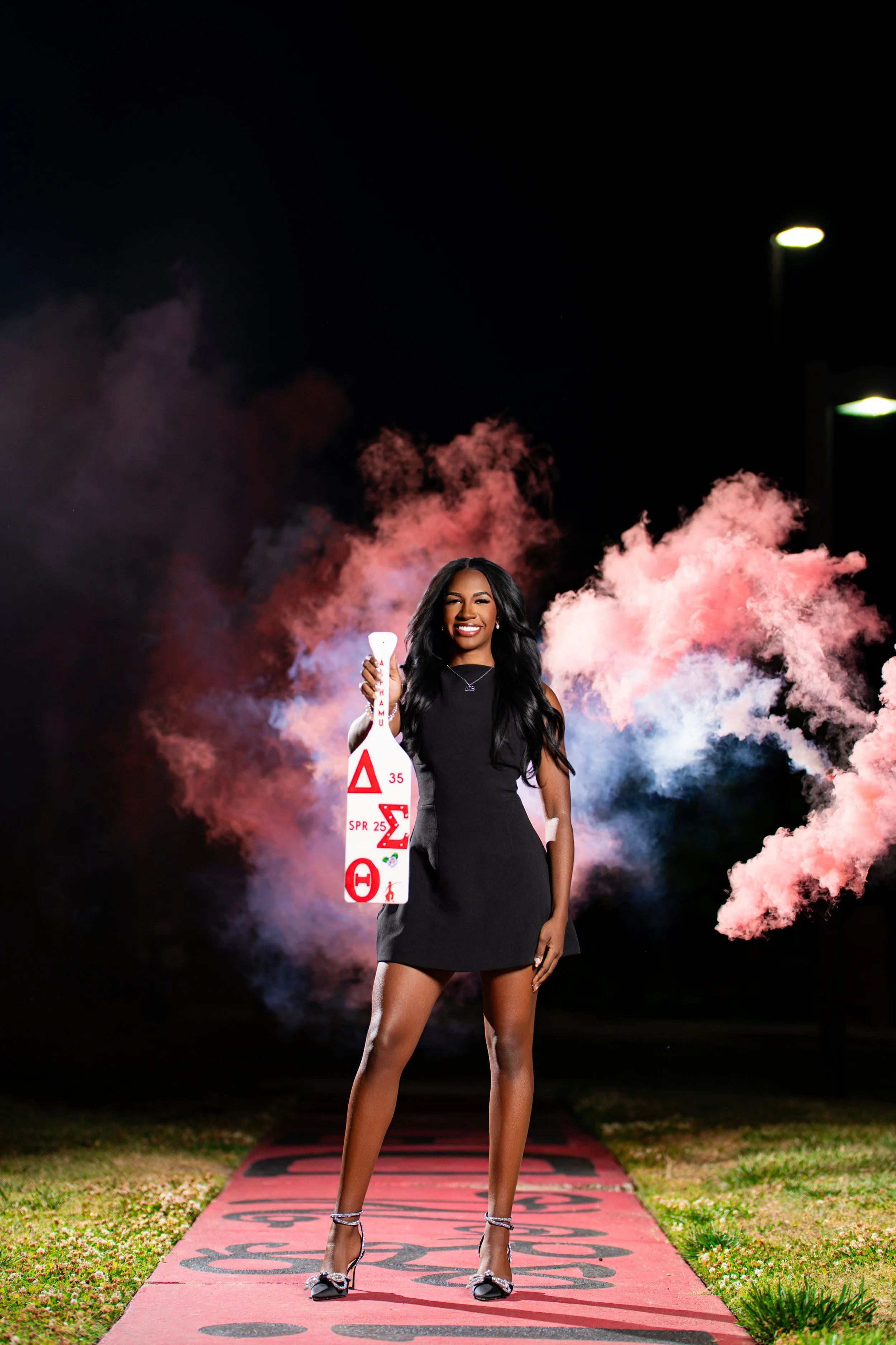 A woman in a black dress stands on a red carpet outdoors at night, holding a white bottle with red labels. Pink smoke clouds glow behind her.