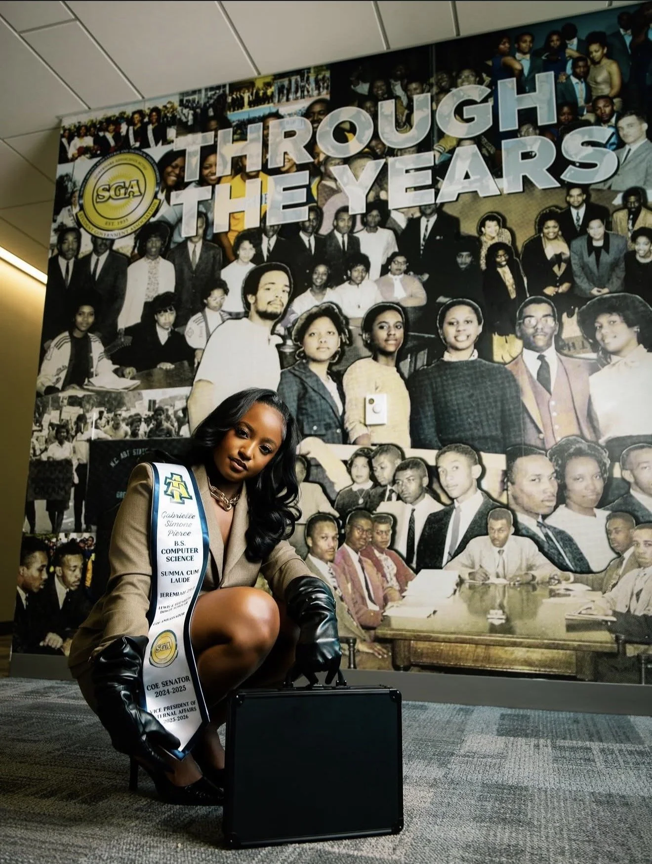 A woman with long dark hair, wearing a beige blazer, black gloves, and a sash, is kneeling in front of a large collage of historical photographs honoring the history of the Sigma Gamma Rho sorority. The sash reads "Gabrielle Simone Pierce, B.S. Compu