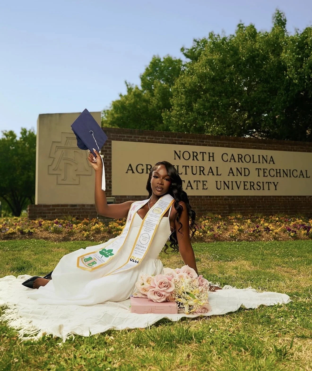 A young woman wearing a graduation cap and gown, seated on a white blanket decorated with pink roses, celebrating her graduation outside in front of a university sign that reads 'North Carolina Agricultural and Technical State University.' She holds 
