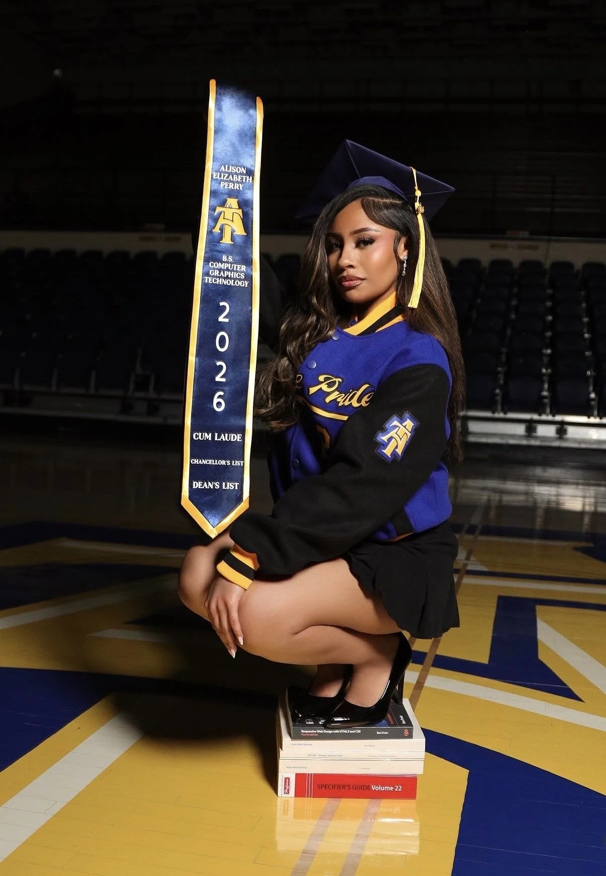 A woman in a graduation cap and gown kneeling on a stack of textbooks on a basketball court, holding a blue and yellow graduation sash with her name and degree, in a sports arena.