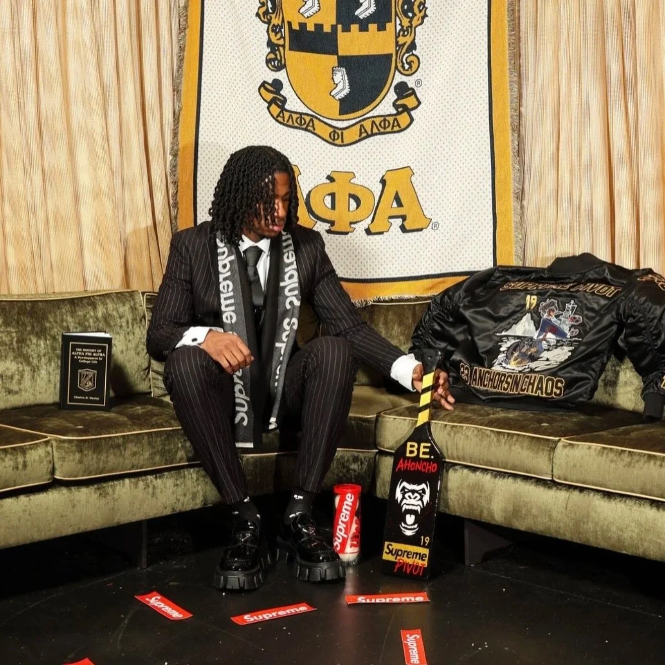 A man dressed in a pinstripe suit with a Supreme scarf, sitting on a vintage green velvet sofa. Behind him is a large Alpha Phi Alpha fraternity banner, with a black and gold satin jacket and various Supreme-branded items displayed on the sofa and floor, including a skateboard, a water bottle, and stickers. The setting appears to be a celebration or fraternity event.
