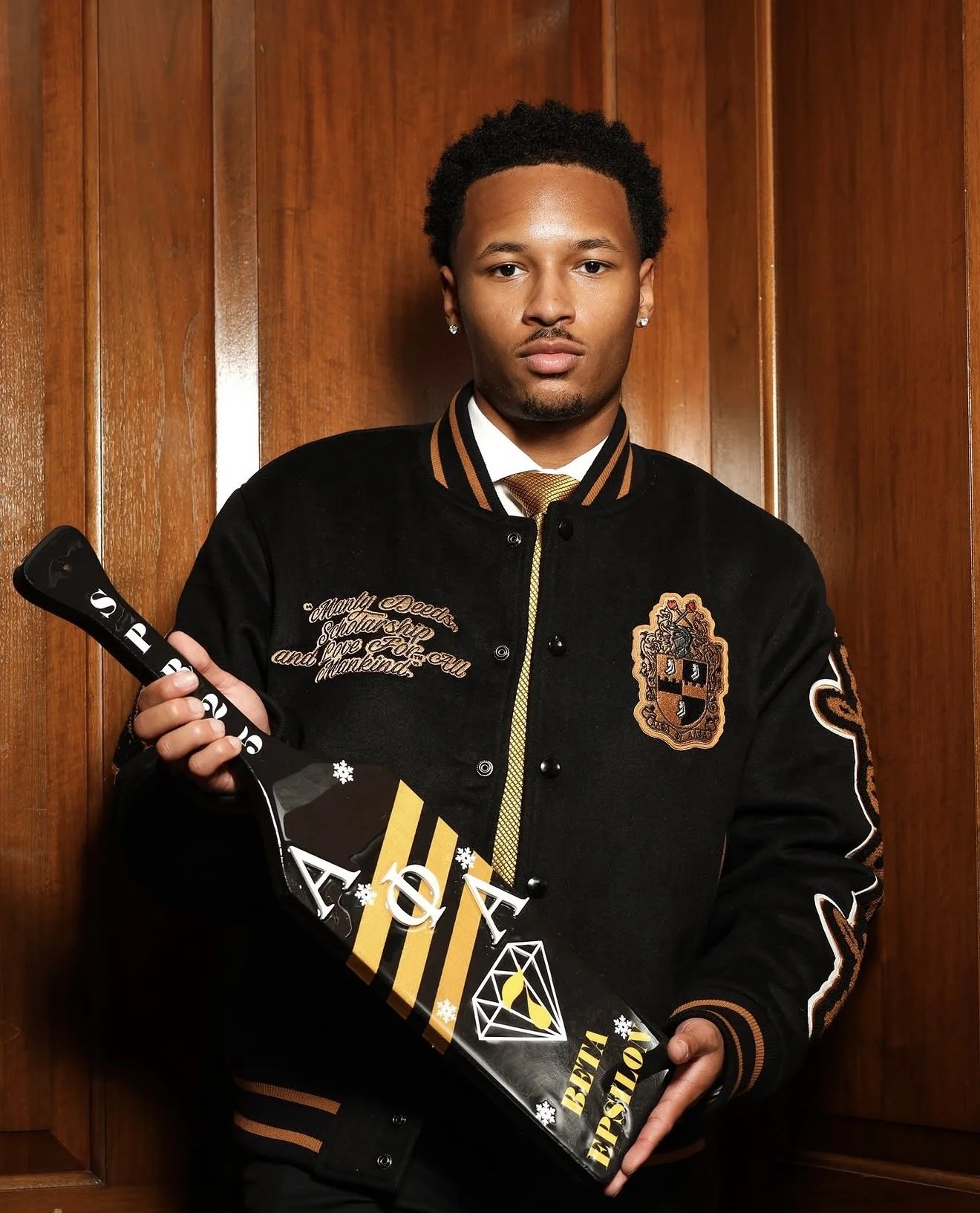 A young man with short curly hair and earrings standing against a wooden wall, holding a black and yellow hockey stick with white snowflake patterns and a diamond logo.