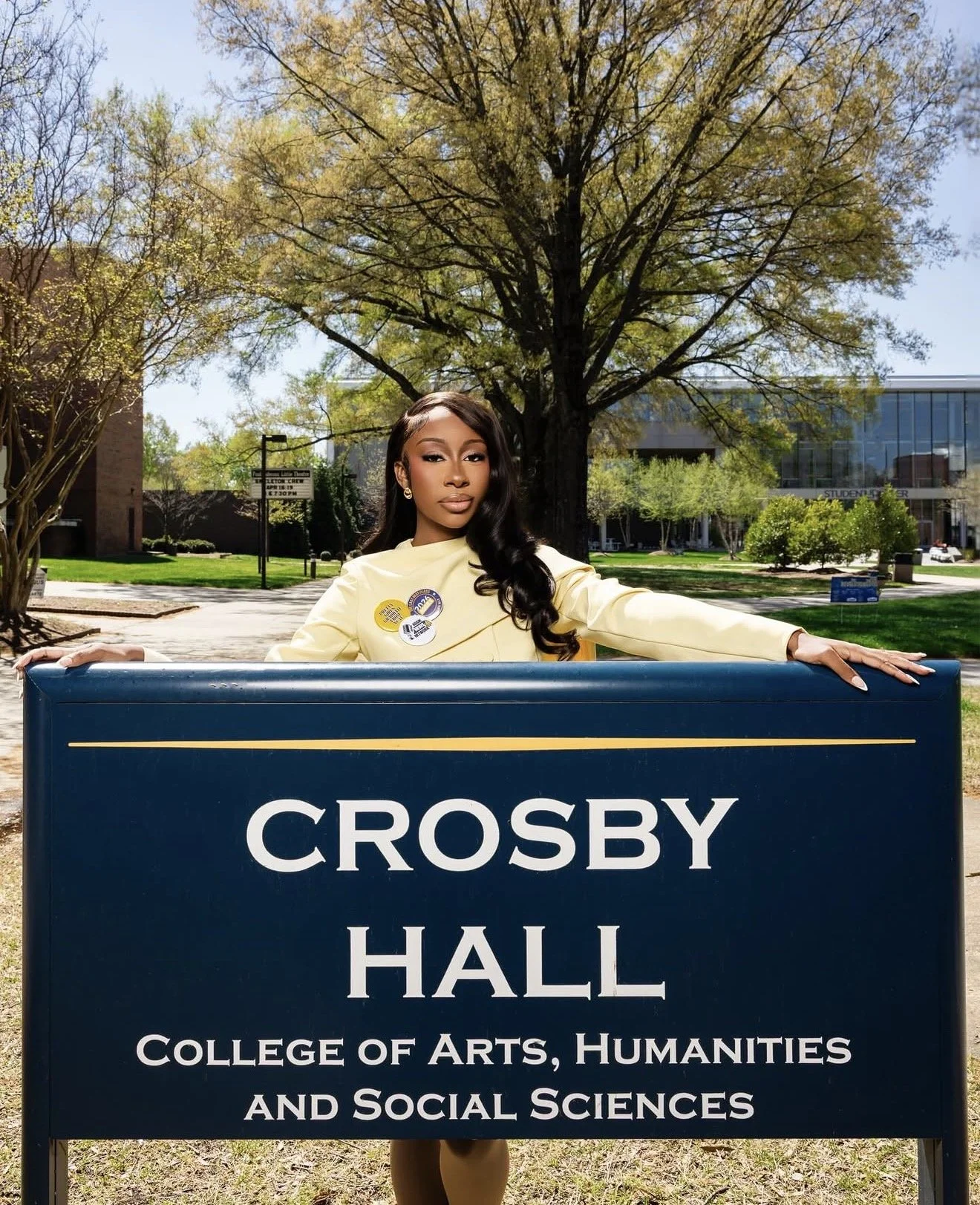 A woman standing behind a sign that reads 'Crosby Hall, College of Arts, Humanities, and Social Sciences' on a college campus with a large tree and buildings in the background.
