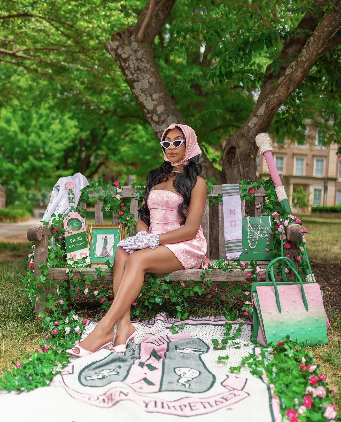 A woman dressed in pink vintage-style dress and matching headscarf, sitting on a decorated wooden bench in a park. She wears white gloves and white high-heeled shoes, with sunglasses on. The bench is adorned with pink flowers and greenery, and surrounded by various fashion accessories, bags, jewelry, and scarves. There is a towel or blanket with graphics and text spread on the ground in front of her, and the background features lush green trees and a building.